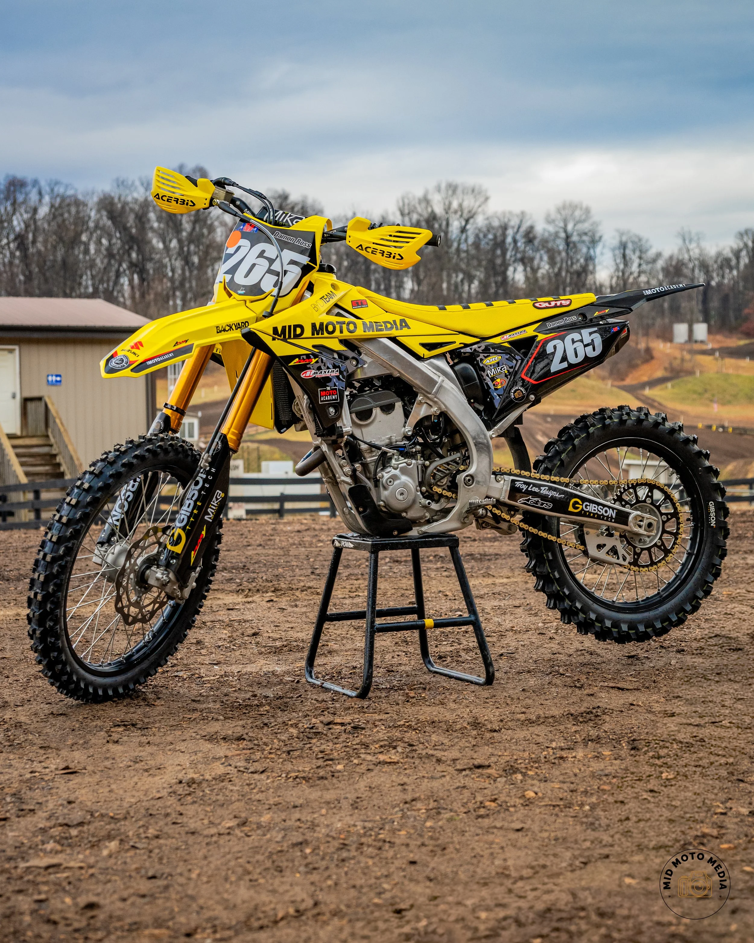 Yellow motocross dirt bike with the number 265 on the front and side, standing on a black stand on a dirt track with trees and a small building in the background.