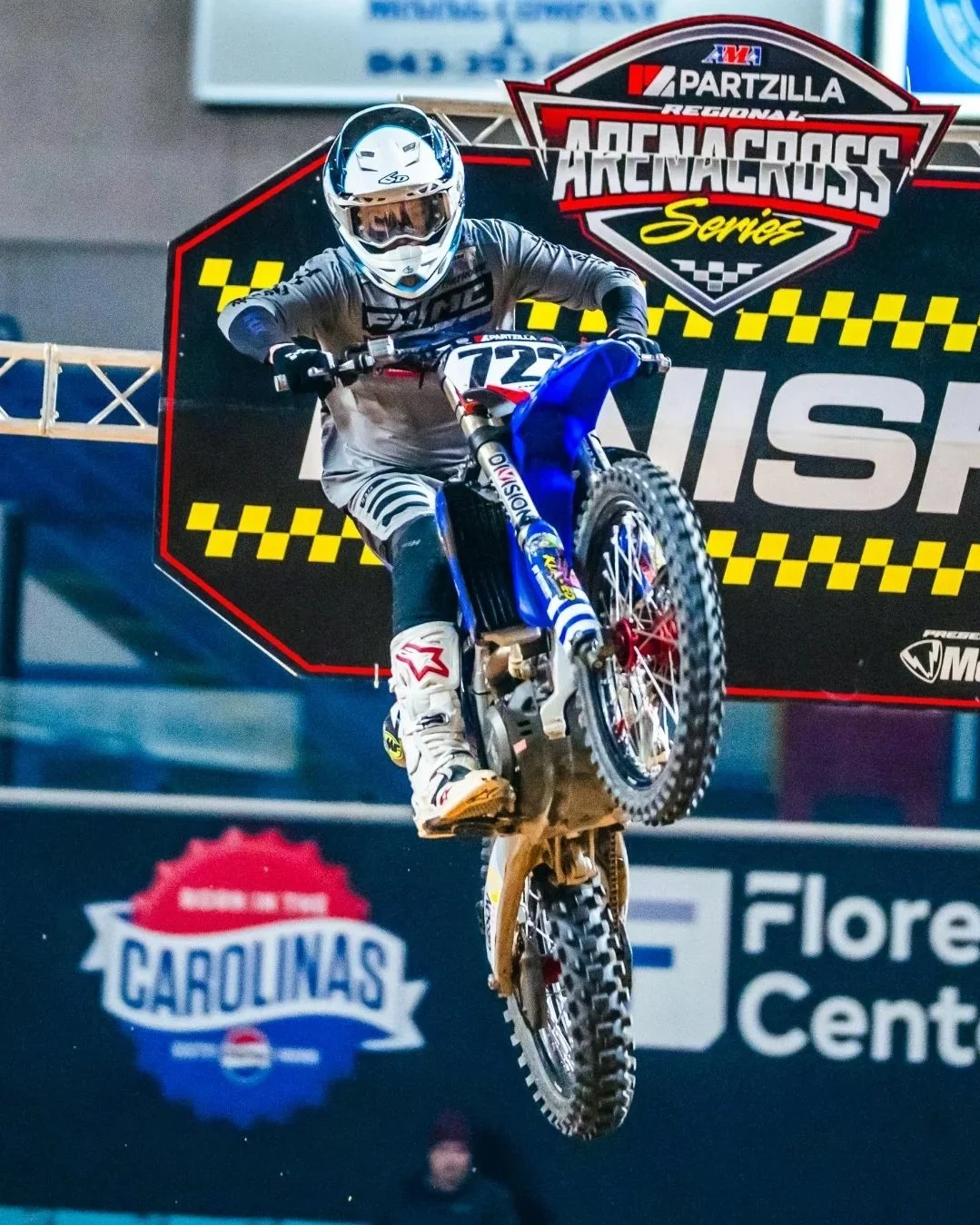 Motocross rider mid-air during a race at the Partzilla Arenacross Series, with a large sign showing the logo and stadium of spectators behind.