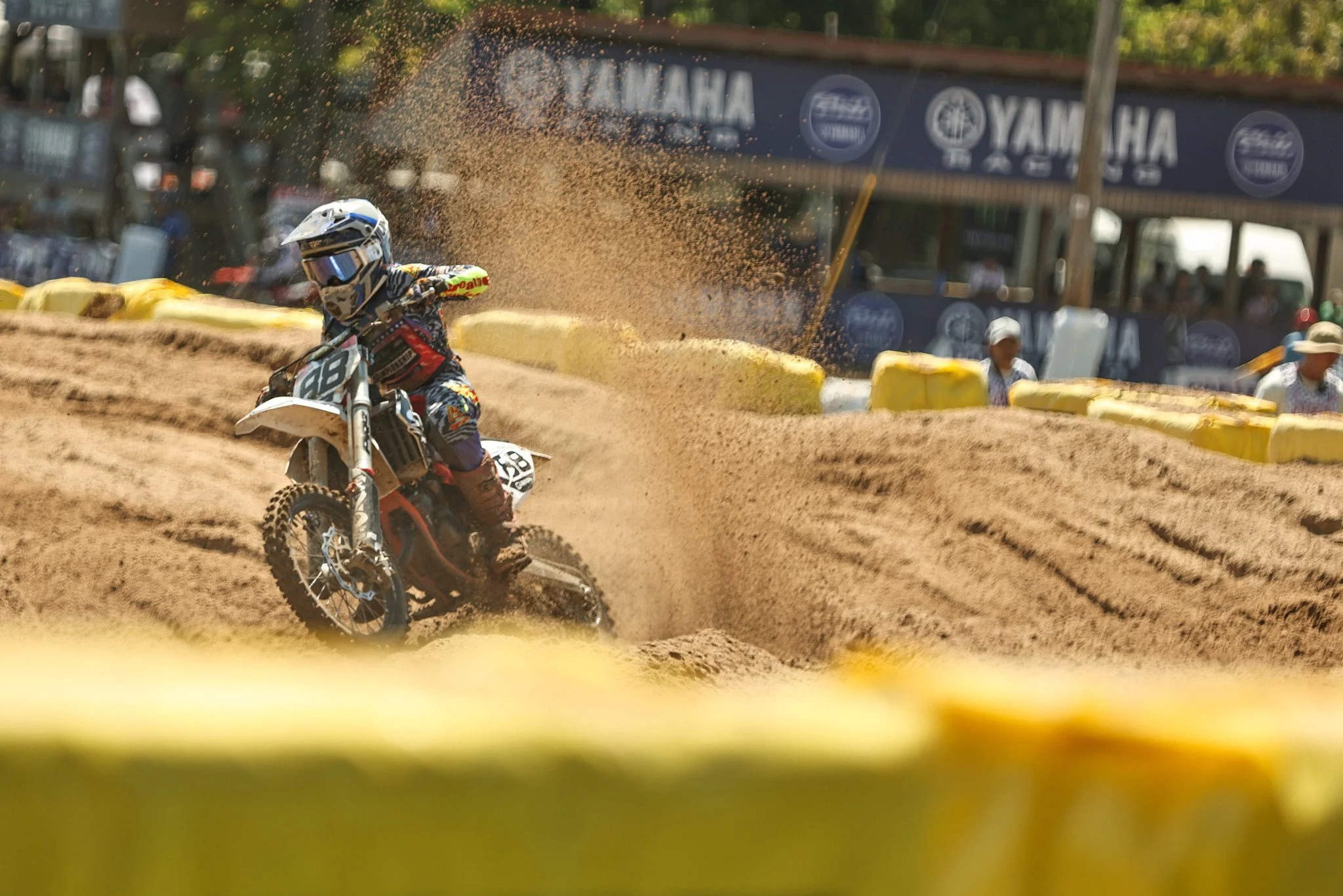 A motocross rider wearing a helmet and colorful gear riding a dirt bike on a dirt track, with dust and dirt flying behind.