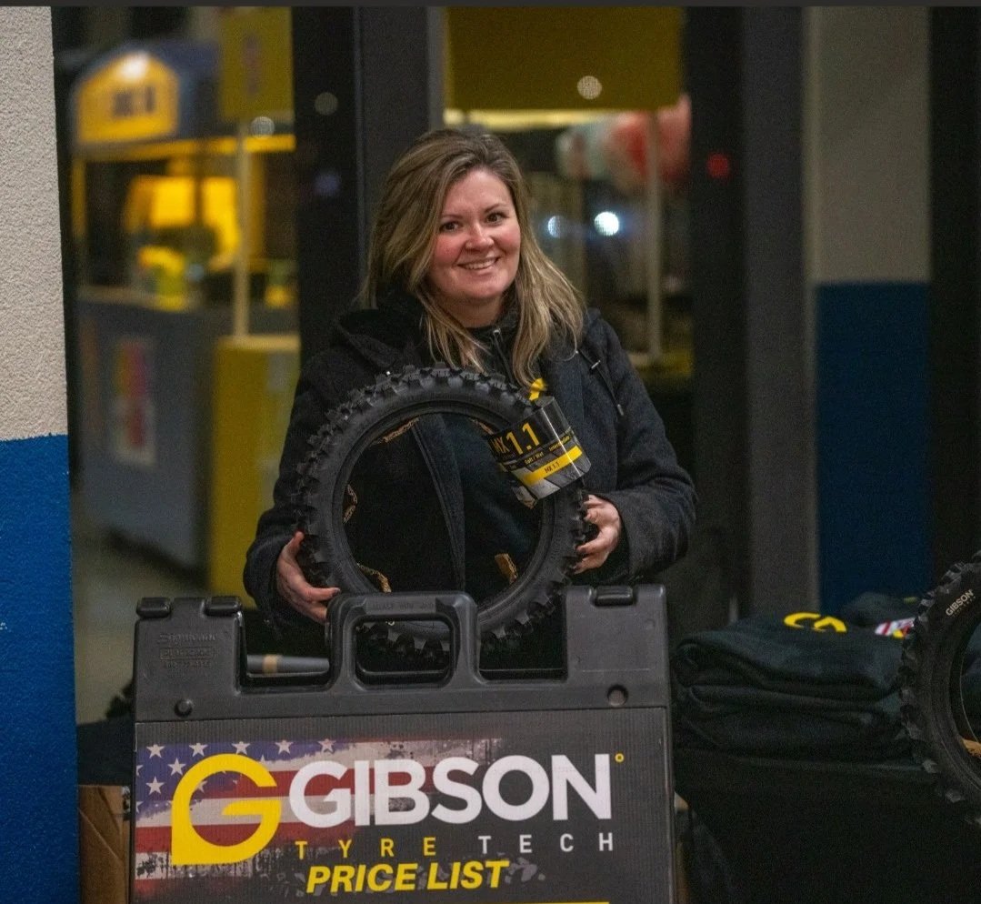 A woman with shoulder-length hair smiling at an indoor race event, holding a small dirt bike tire with a Gibson Tire Tech price list sign in front.