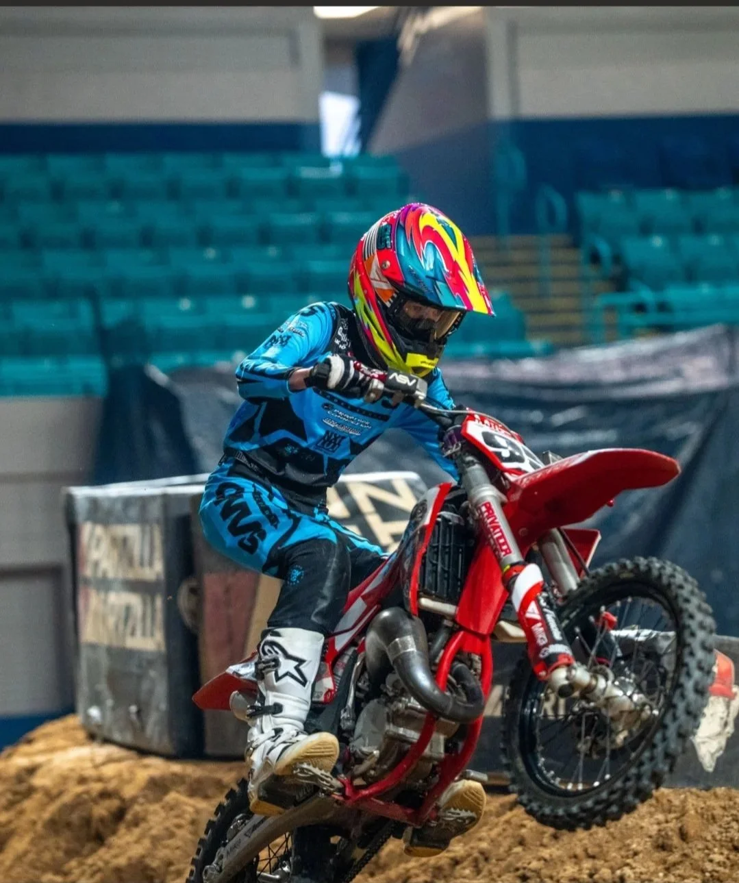A person in motocross gear riding a red dirt bike on an indoor dirt track.
