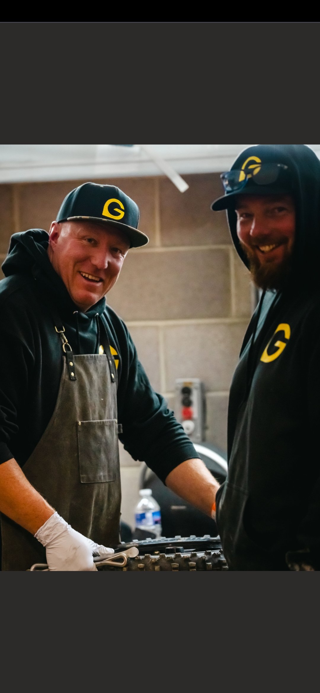 Two smiling men wearing black hoodies with yellow G logos, black caps with yellow G logos, and aprons, working together in an indoor setting with a concrete block wall. One man is wearing a glove and holding tools, possibly working on mechanical or technical equipment.