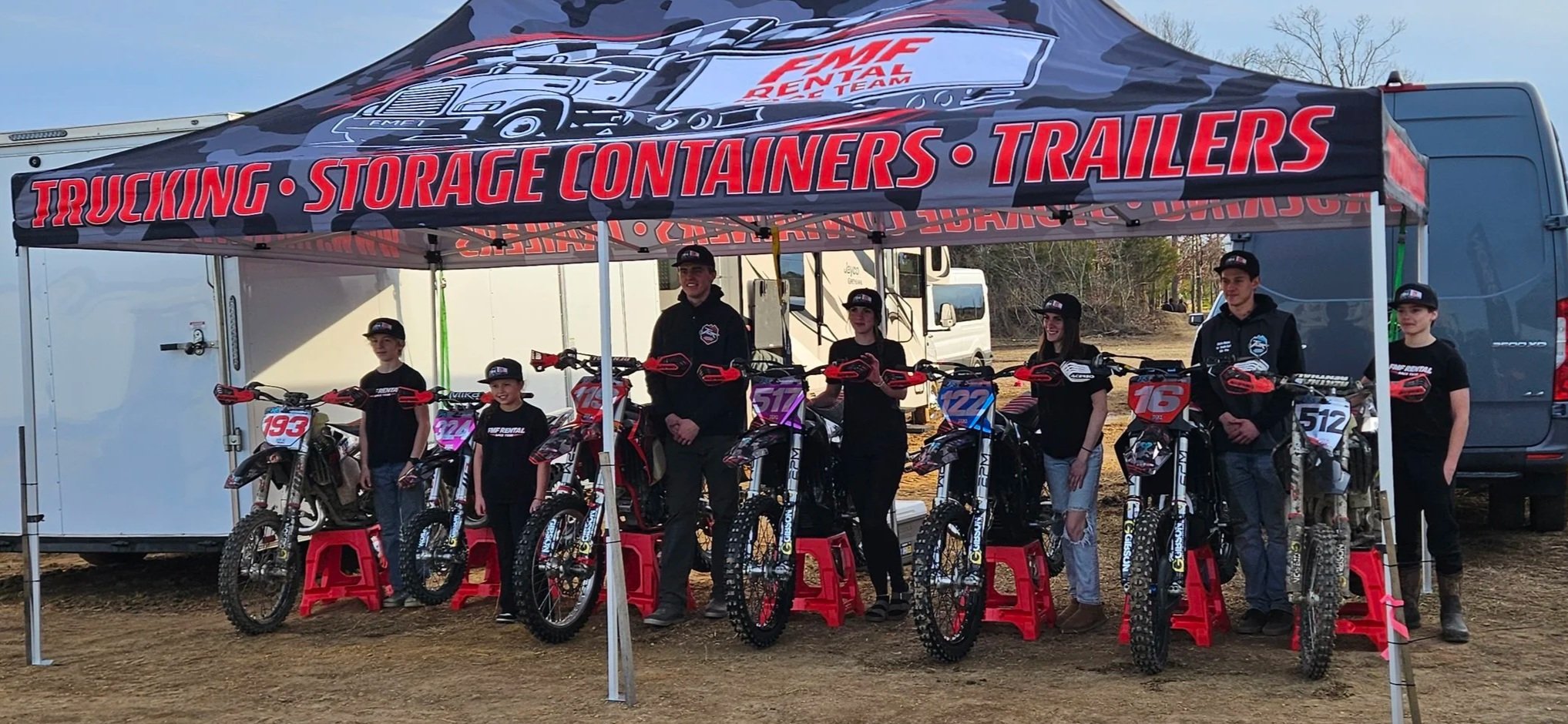 Group of kids and adults with dirt bikes under a canopy that says 'Trucking - Storage Containers - Trailers' at an outdoor dirt bike track.