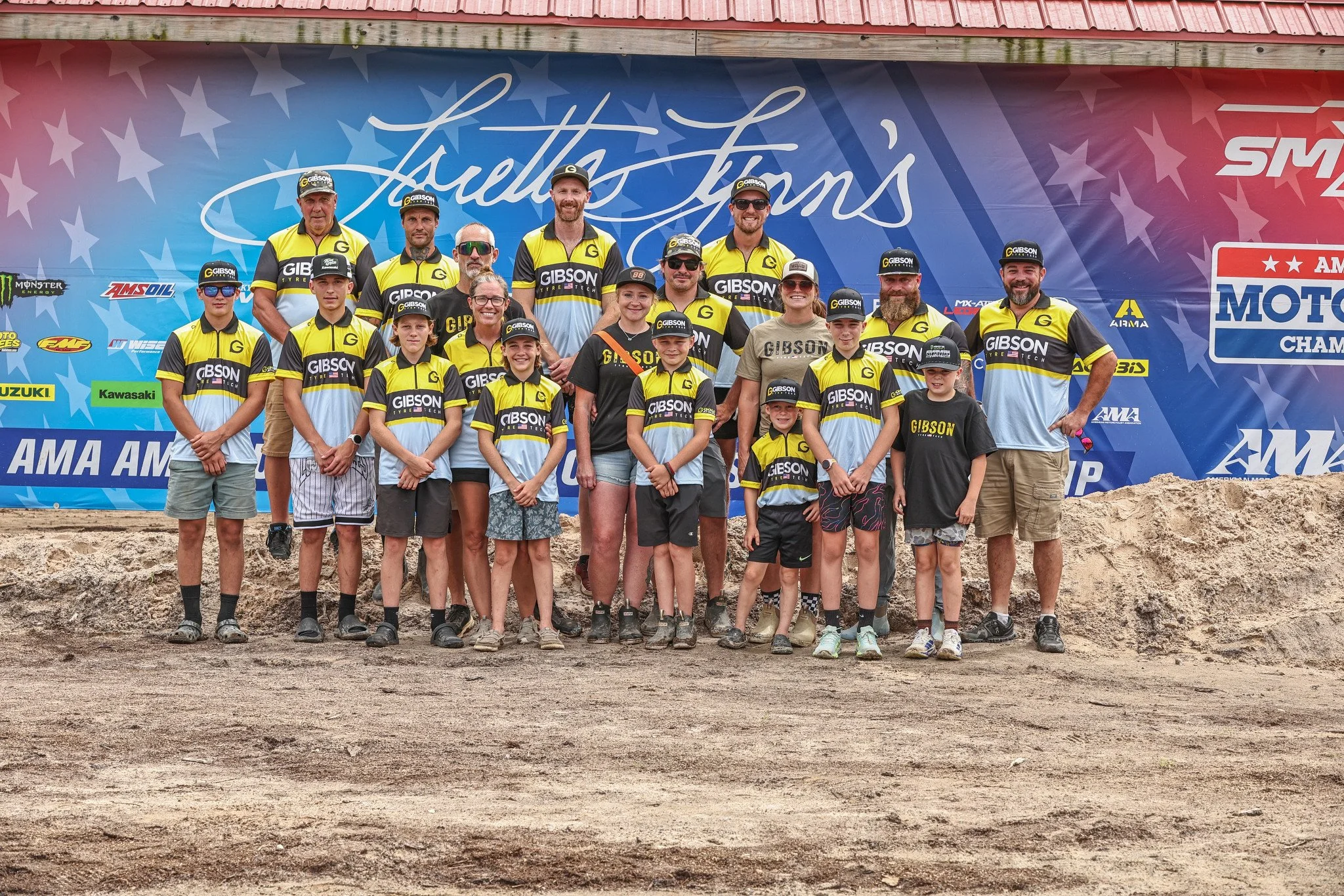 A group of people, including children and adults, standing on a dirt track in front of a blue backdrop with logo and sponsor decals, wearing matching yellow and black team jerseys, some with caps and sunglasses, posing for a team photo at an outdoor motorsports event.