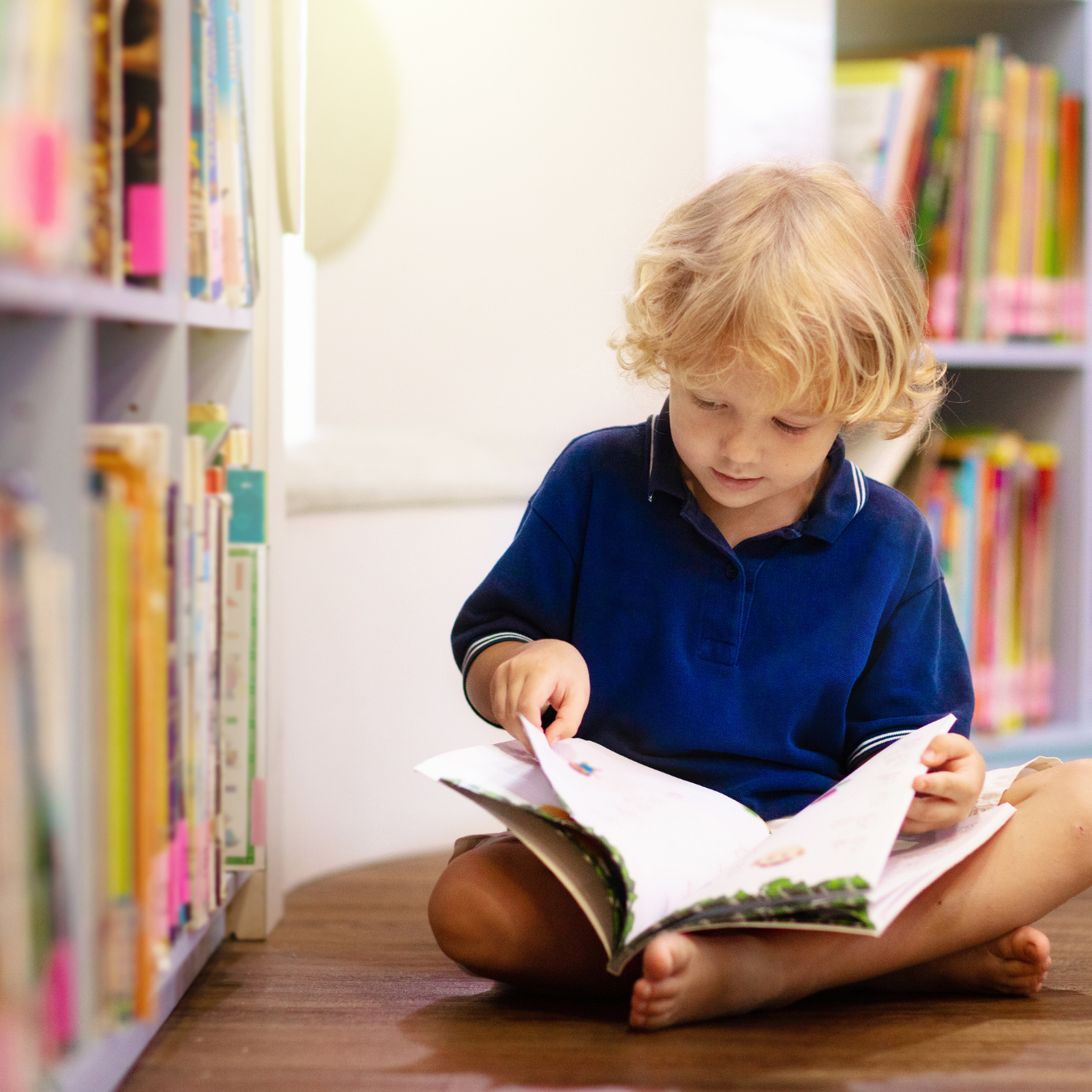 Young student sitting on the floor reading a book beside a classroom bookshelf.