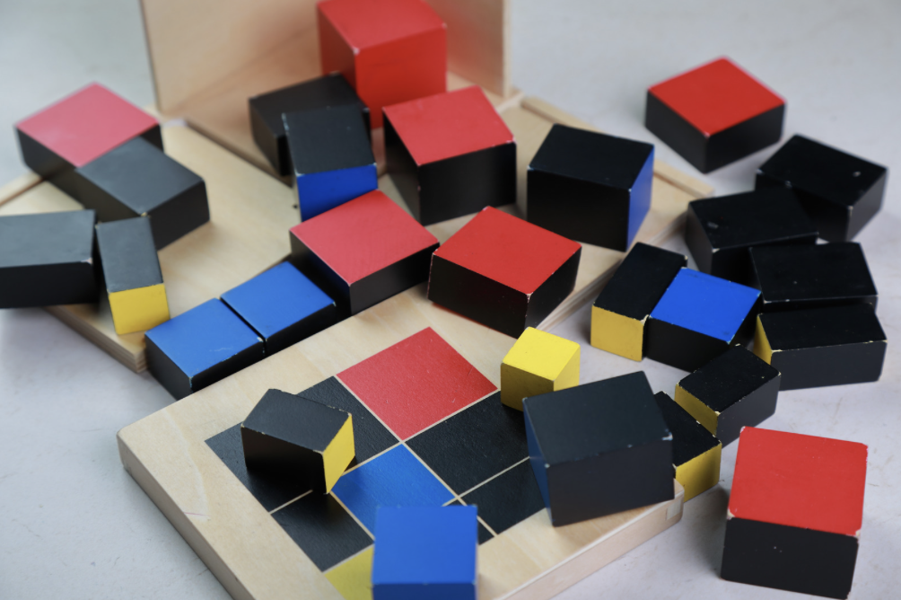 Montessori binomial cube pieces in red, blue, yellow, and black arranged on wooden trays.