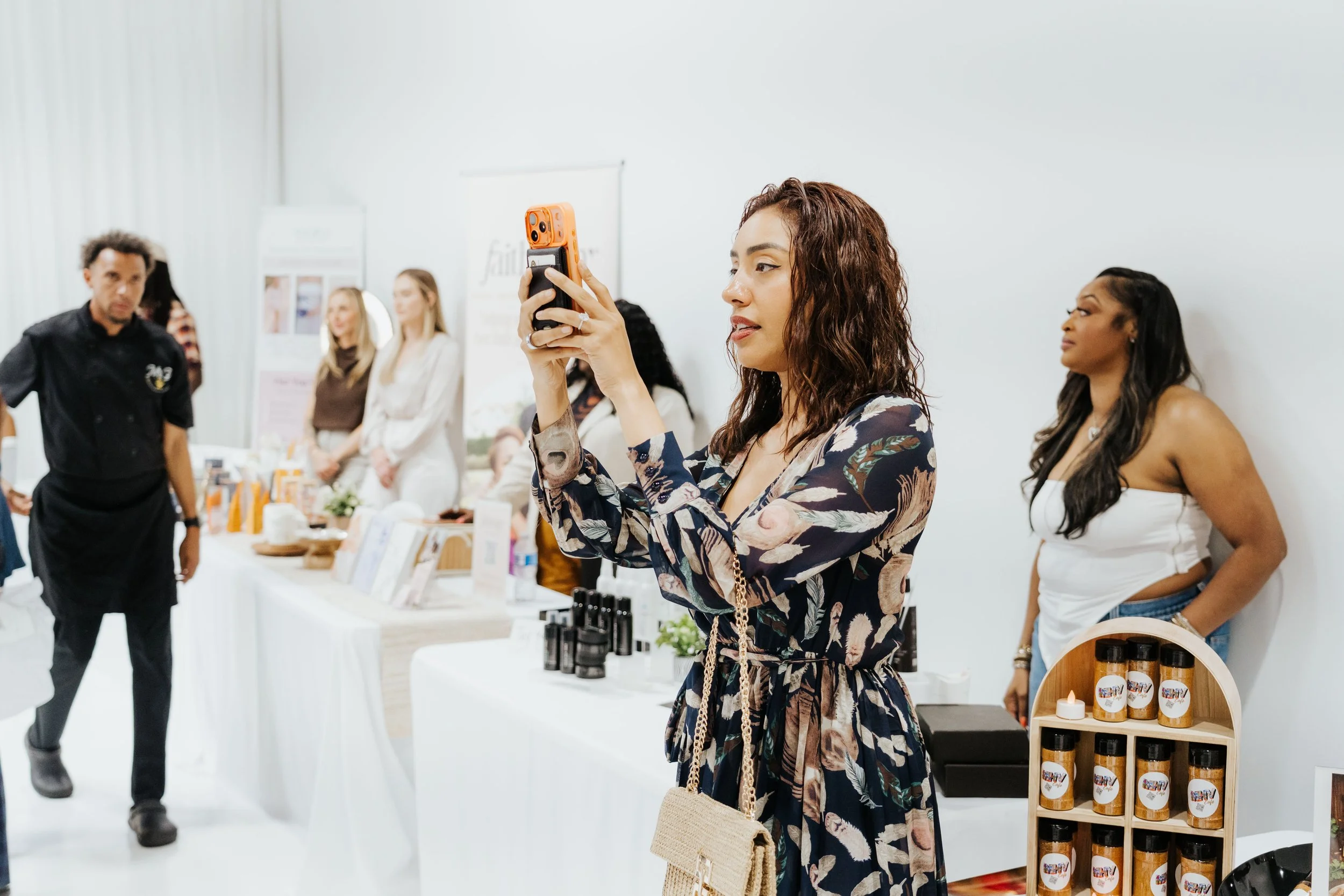 A woman with curly hair taking a photo with her smartphone at an indoor marketplace or event, with other women and tables displaying products in the background.
