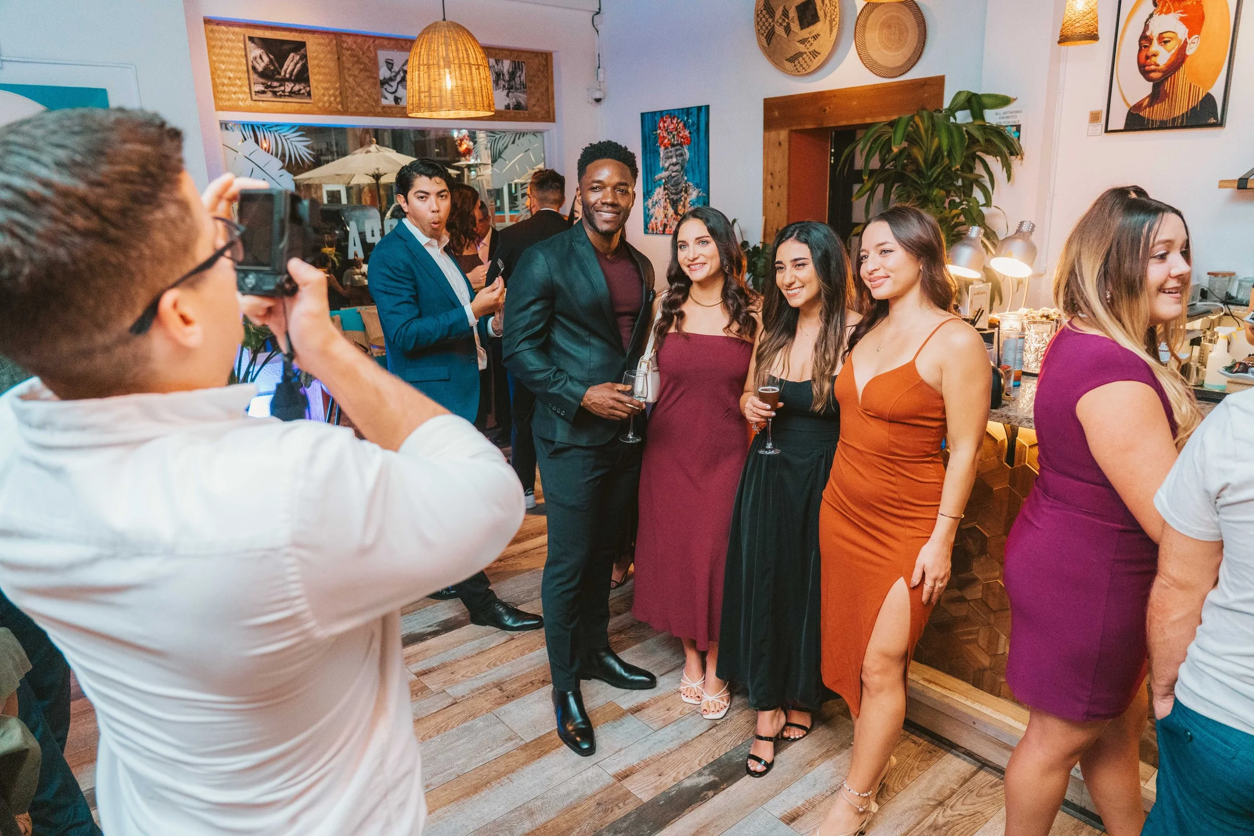 People taking a group photo at a party or celebration, with friends dressed in colorful dresses and suits, in a decorated indoor venue.