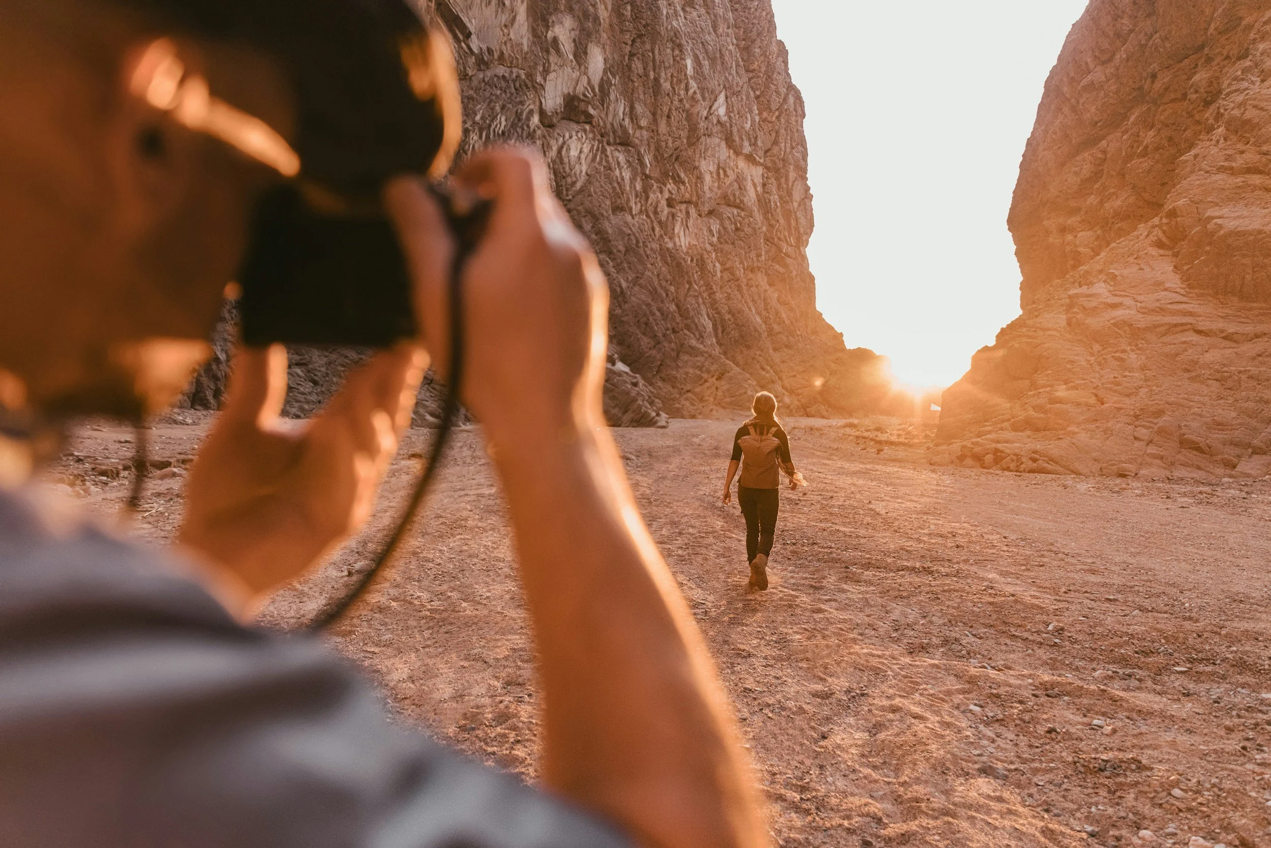 A person taking a photo of another person hiking through a rocky canyon at sunset.