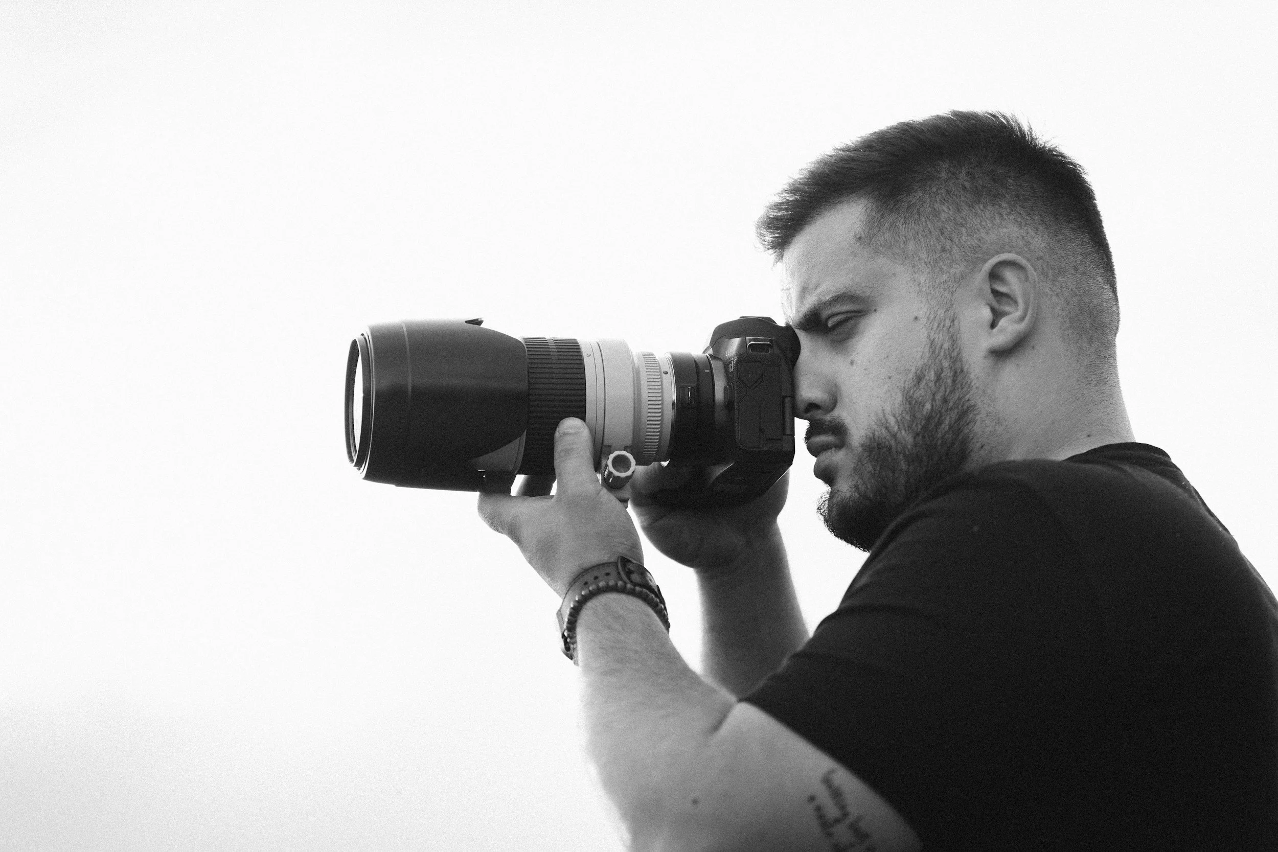A man with short hair and a beard is looking through a camera with a large telephoto lens.