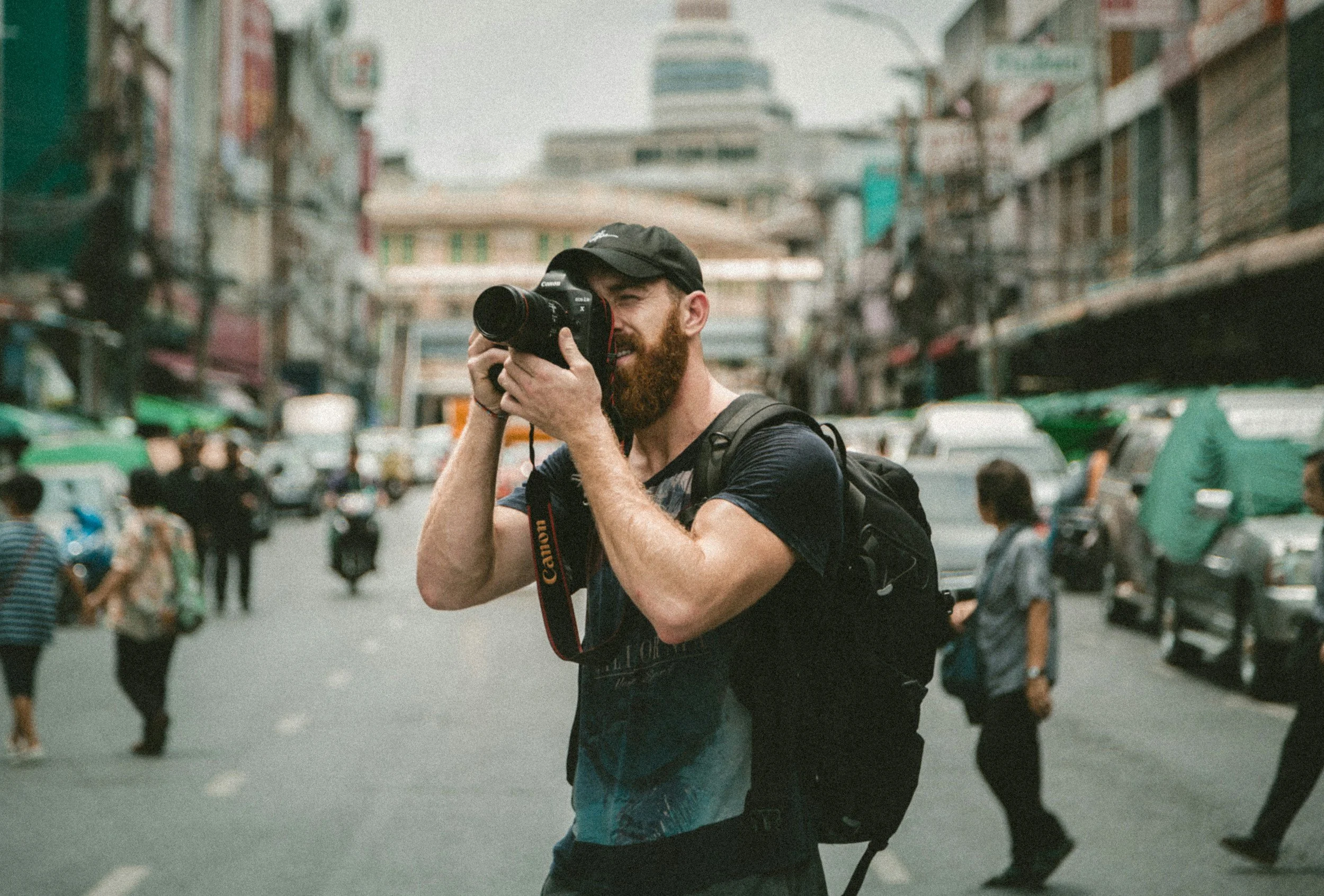 A man with a beard and baseball cap taking a photograph in a busy urban street, carrying a backpack, with pedestrians and vehicles around.