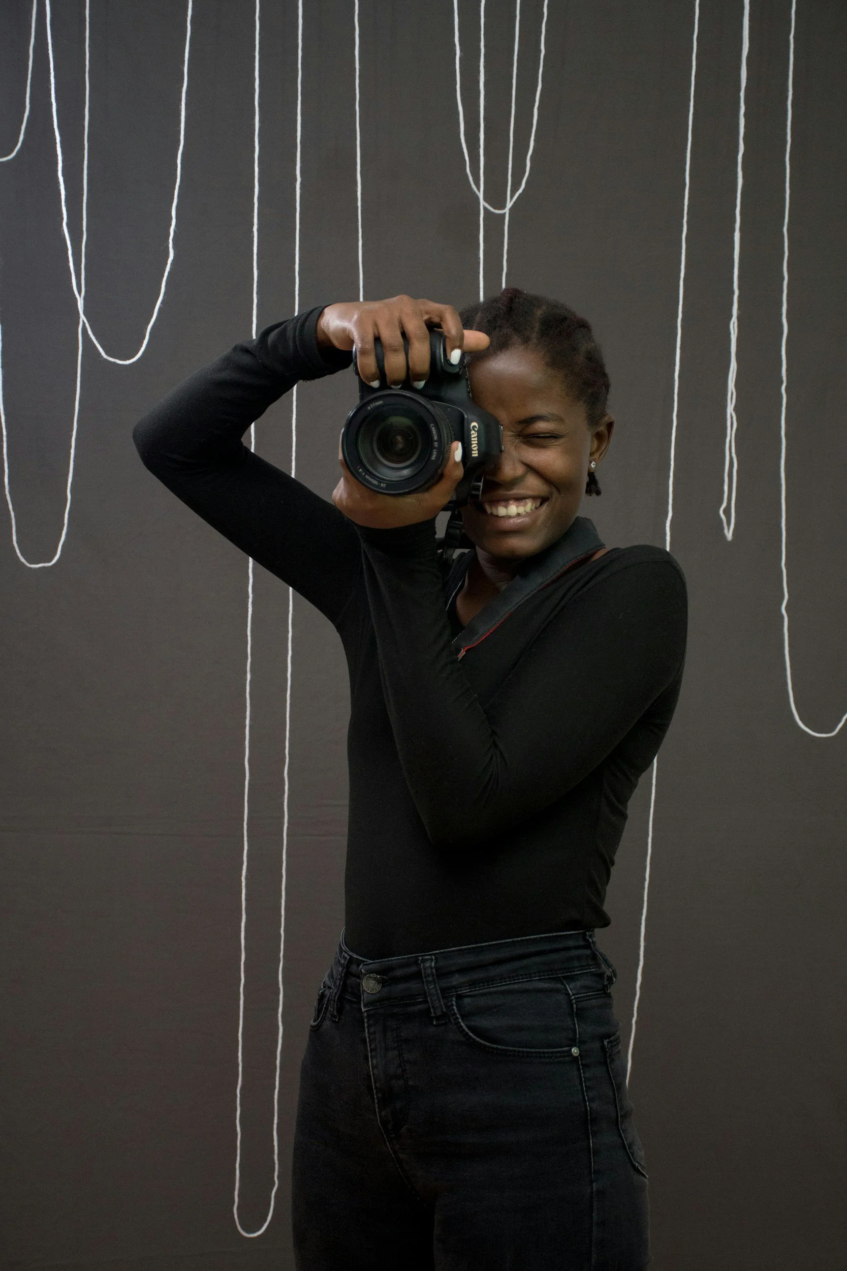 A woman smiles while taking a selfie with a Canon camera in front of a black wall with white string art hanging from the ceiling.