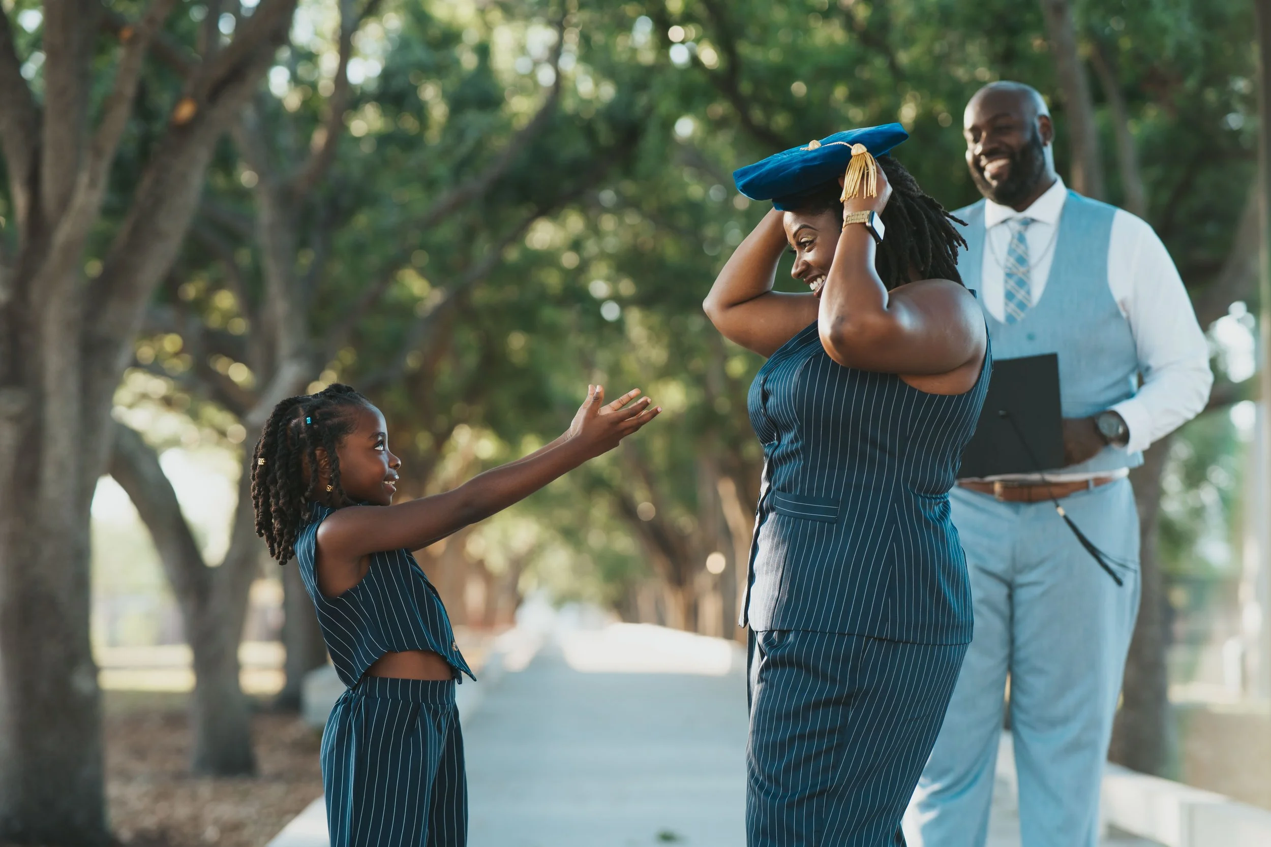 A young girl reaching out to her mother, who is smiling and putting on a graduation cap, while a man in academic attire looks on happily outdoors in a park.