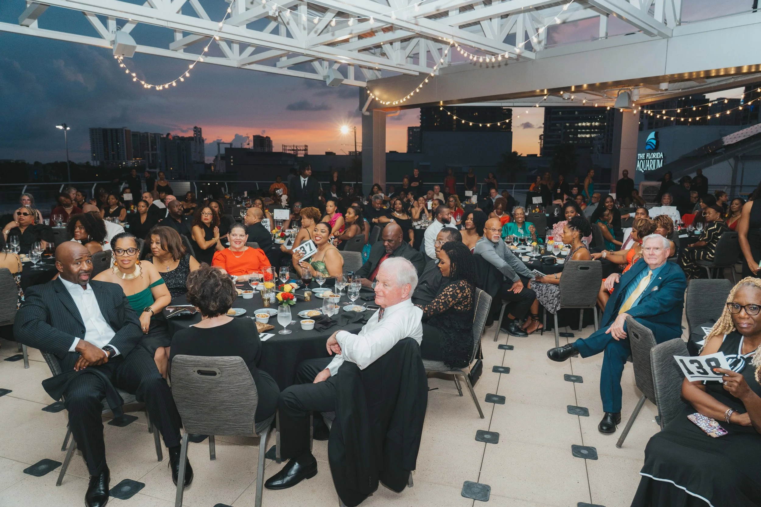 A large group of people sitting at tables on a rooftop during sunset, attending an event or dinner, with city buildings and string lights overhead.