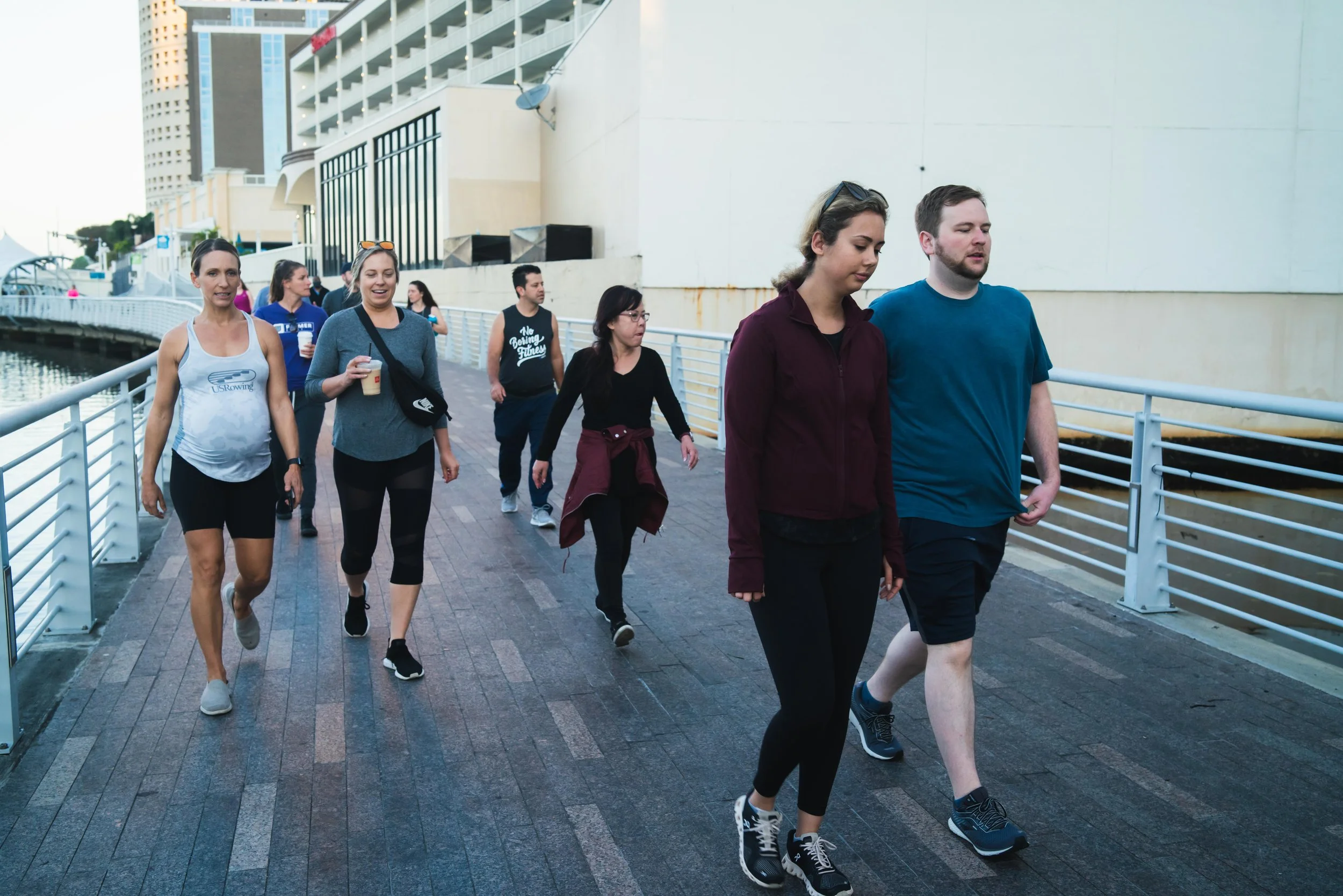 Group of people walking along a waterfront promenade, with modern buildings behind them.
