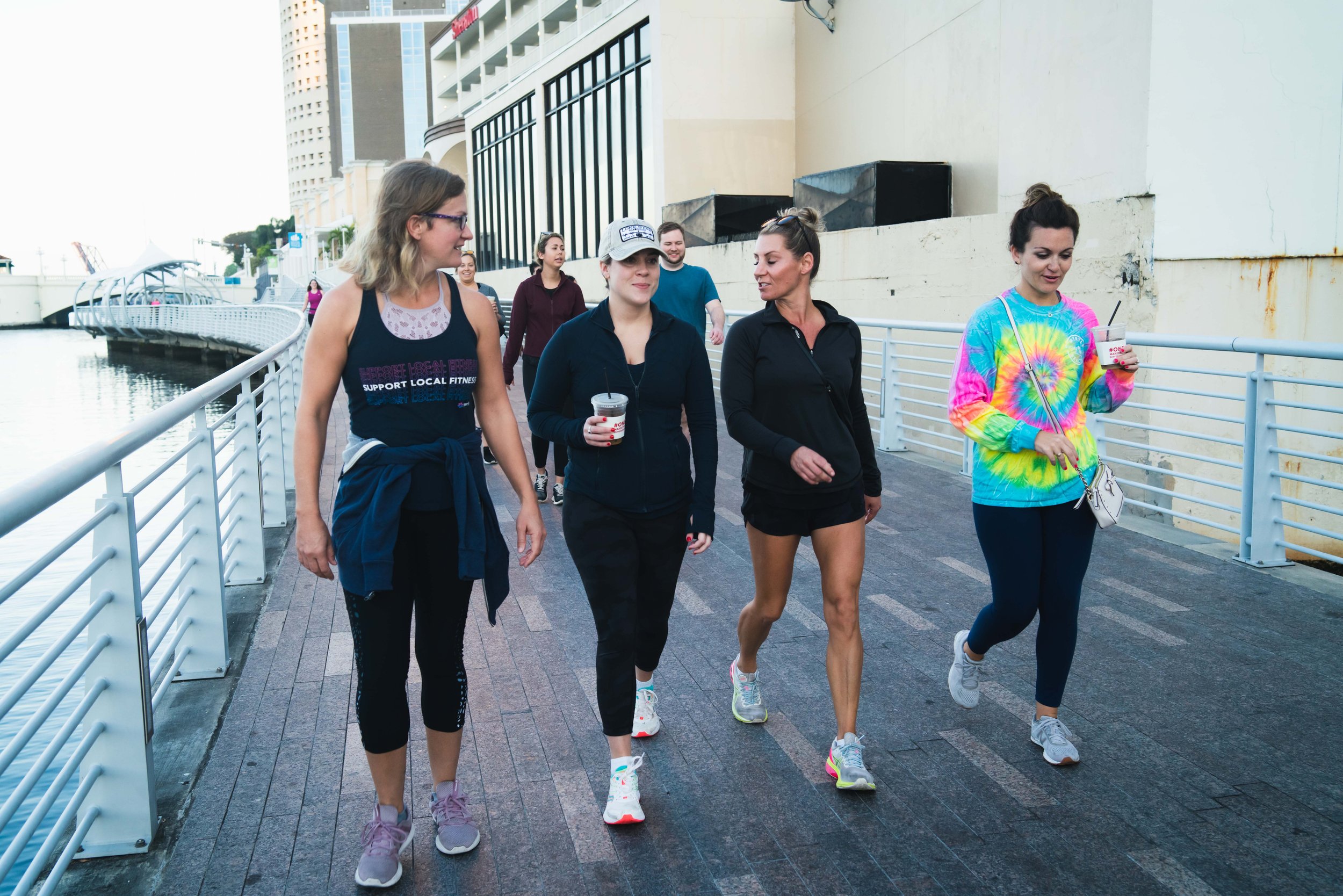 Group of five women walking along a waterfront path, some holding drinks, in athletic clothing, with city buildings in the background.