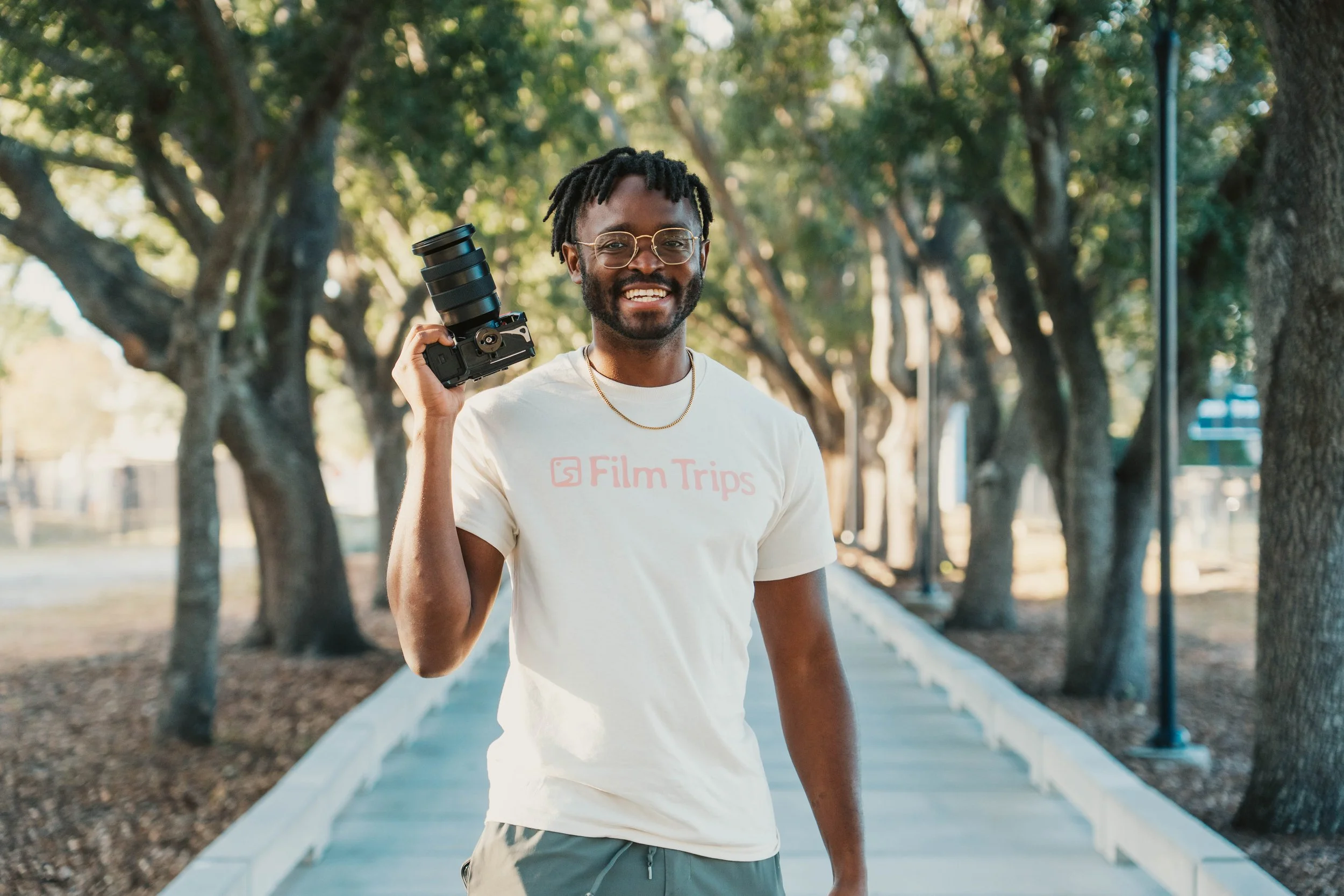 A smiling man with dreadlocks and glasses holding a camera over his shoulder, walking on a tree-lined sidewalk.
