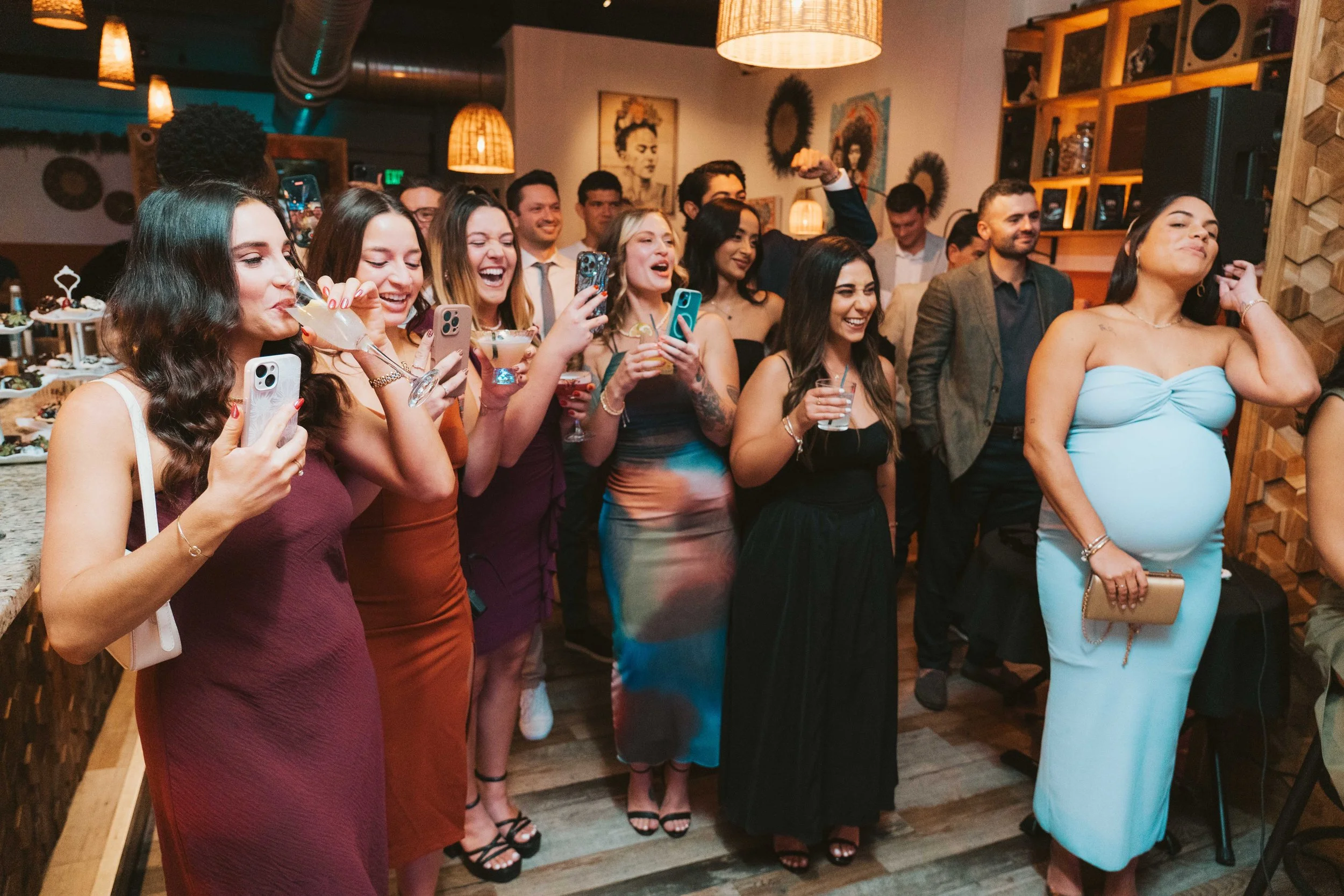 Group of people at a party, smiling, taking photos, and enjoying drinks, in a warmly lit indoor setting with artwork on the walls and a wood shelf.