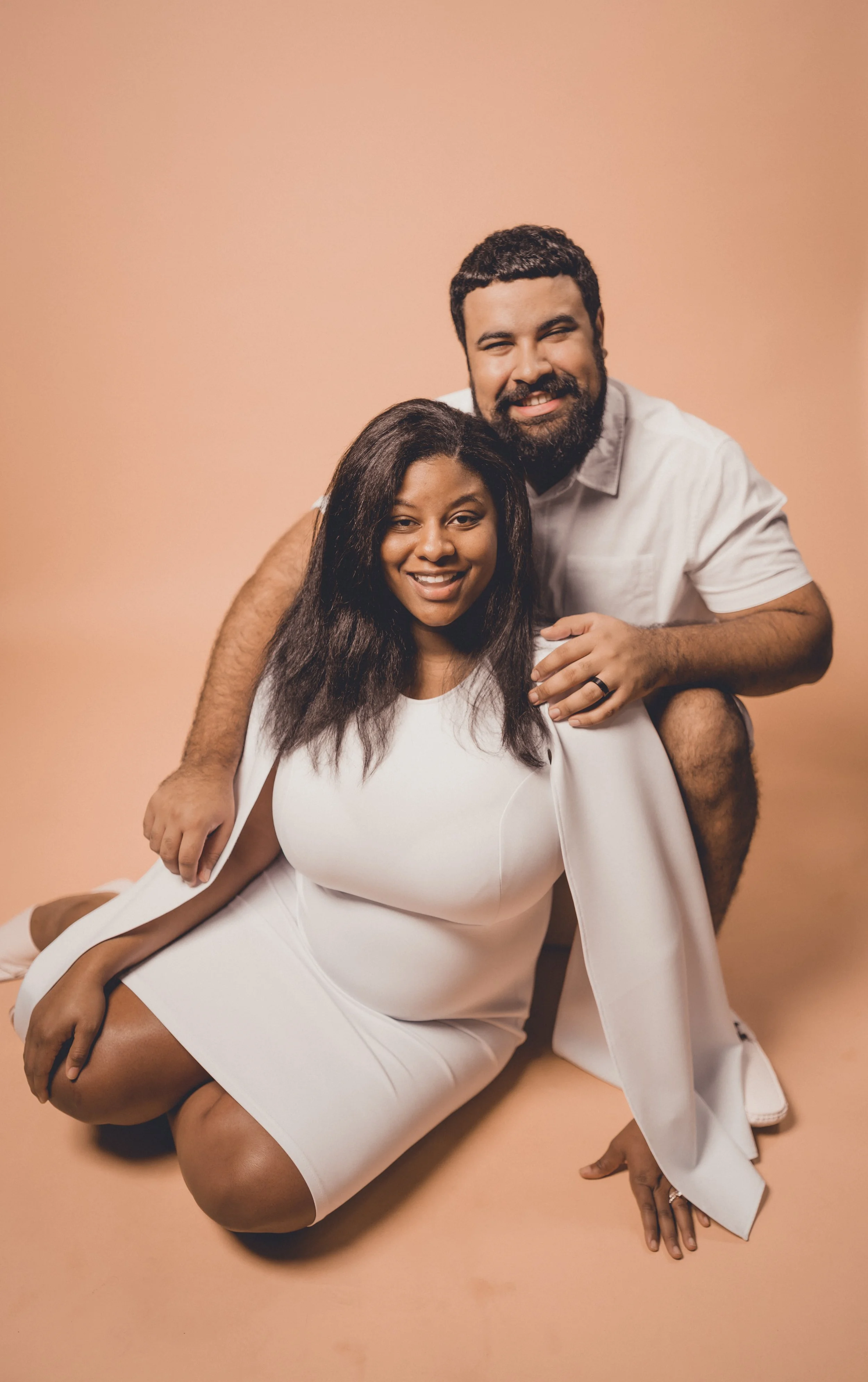 A happy couple poses together against a peach-colored background. The woman is kneeling in front, wearing a white dress, and the man is behind her, leaning on her shoulder, wearing a white shirt. He has a beard and short curly hair, and both are smiling.