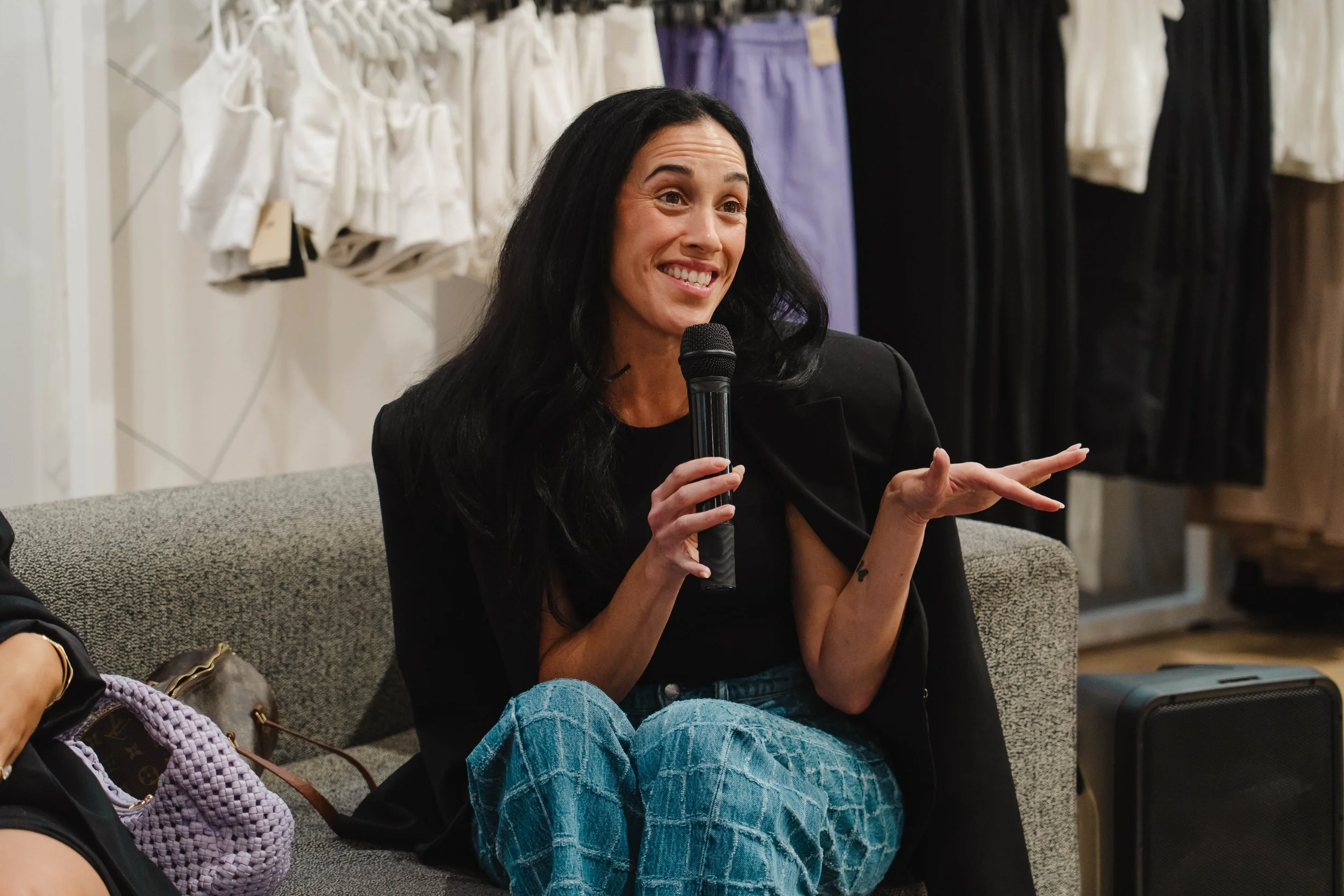 Woman with long dark hair speaking into a microphone during a discussion, sitting on a gray sofa in a room with clothing on display behind her.