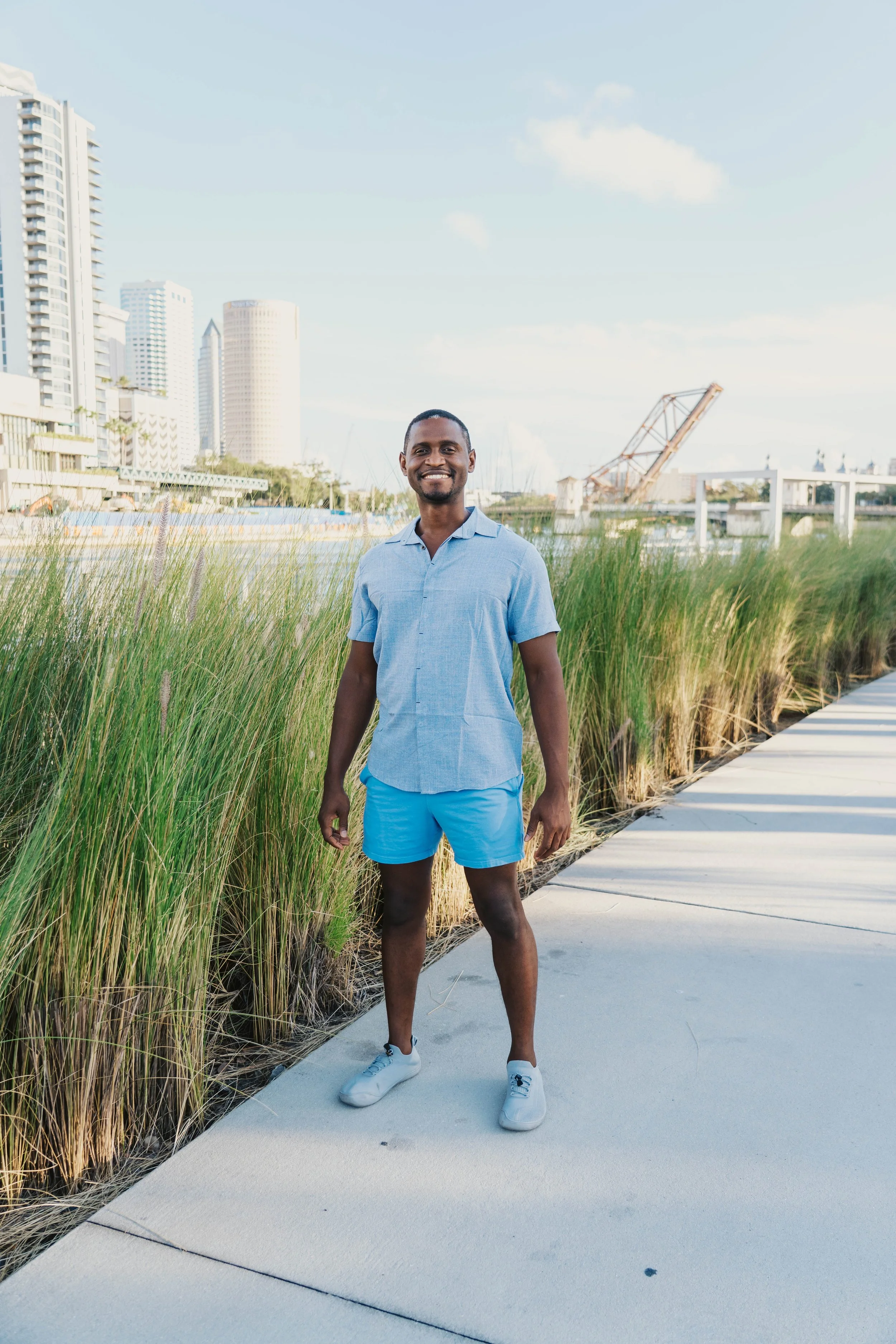 Smiling man in light blue shirt and shorts standing on sidewalk with city skyline and river in background