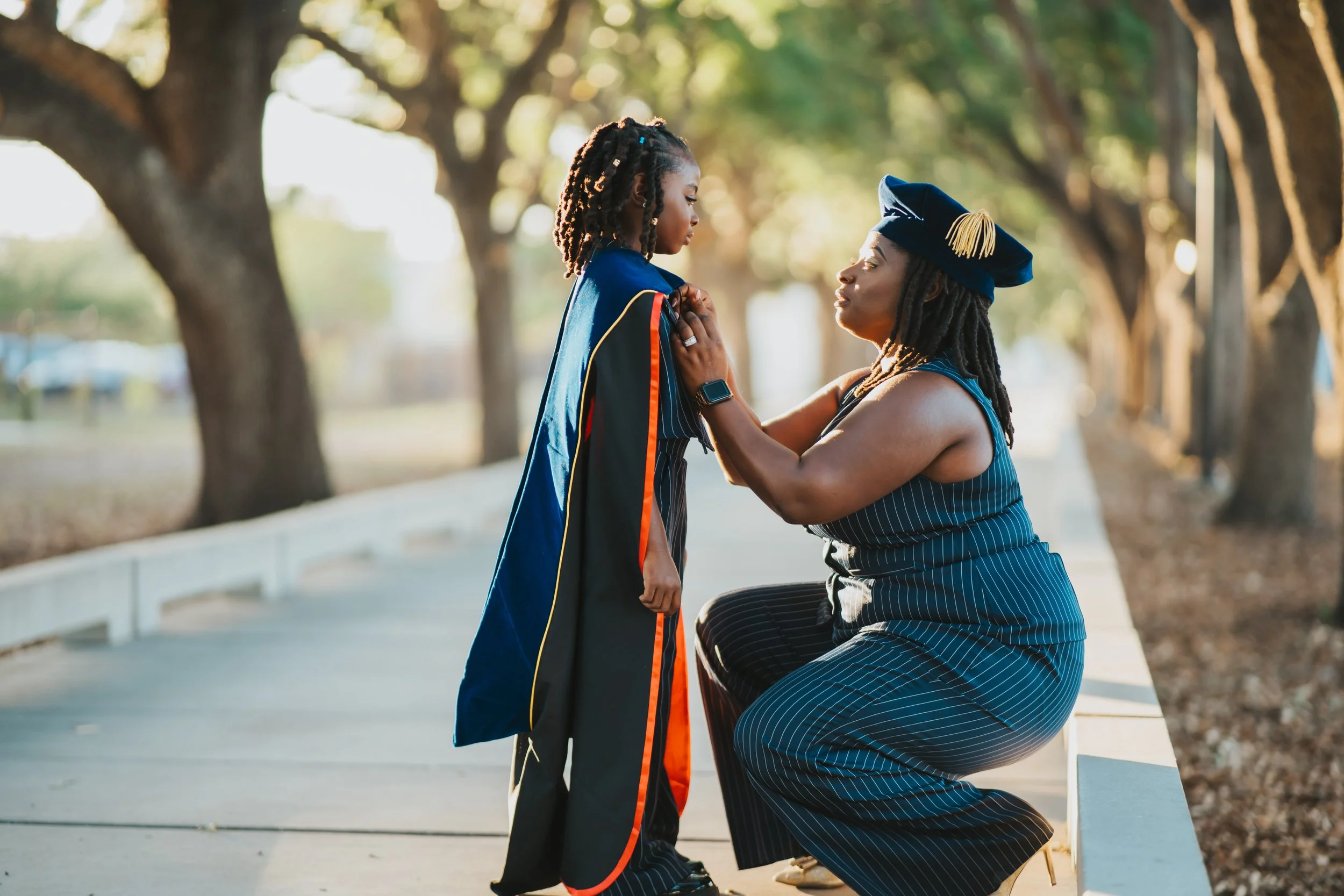 A woman in graduation attire kneeling on a park bench in front of a seated woman, placing a graduation stole around the student's neck during sunset.
