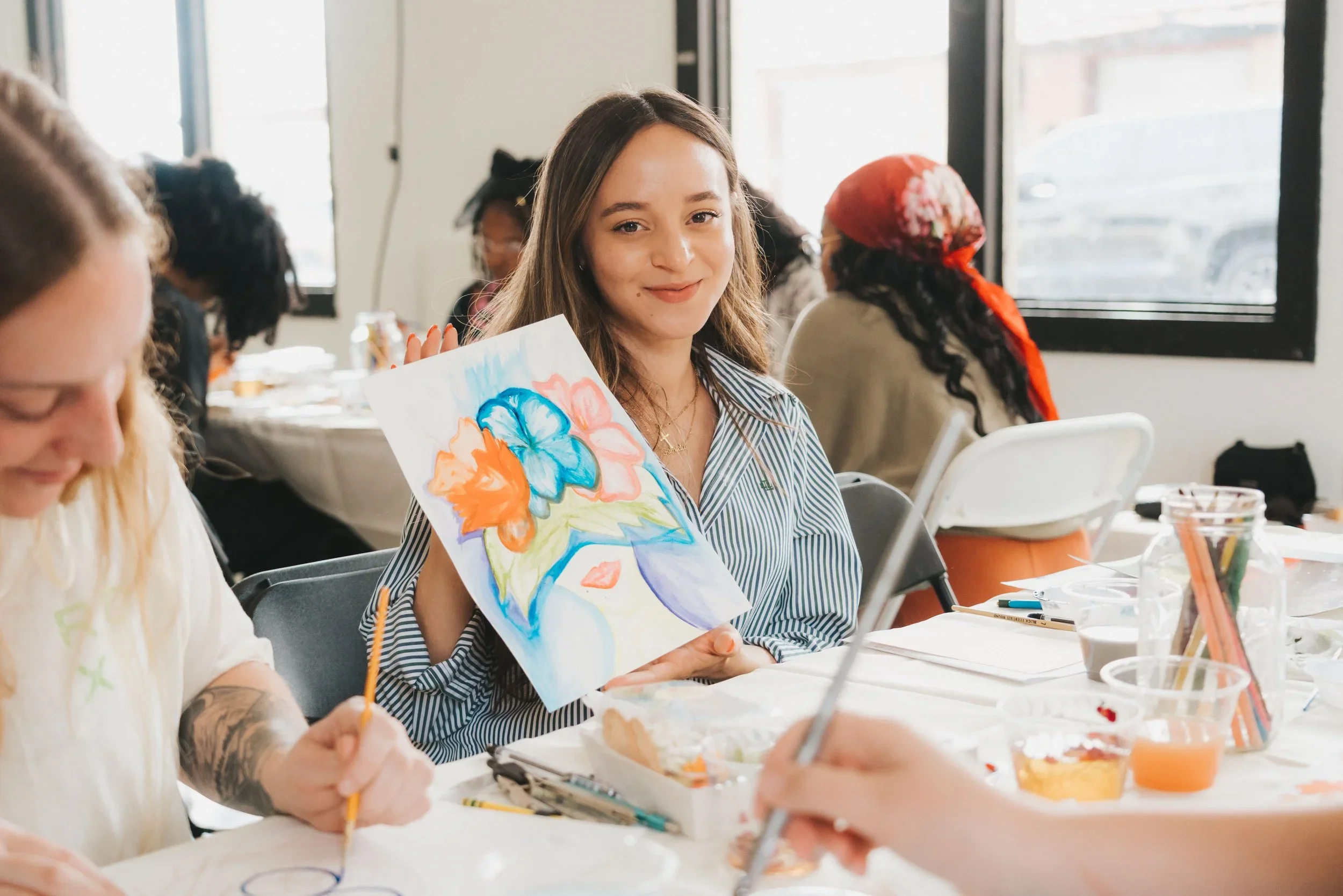 Young woman holding up a colorful floral painting at an art class with others around a table.