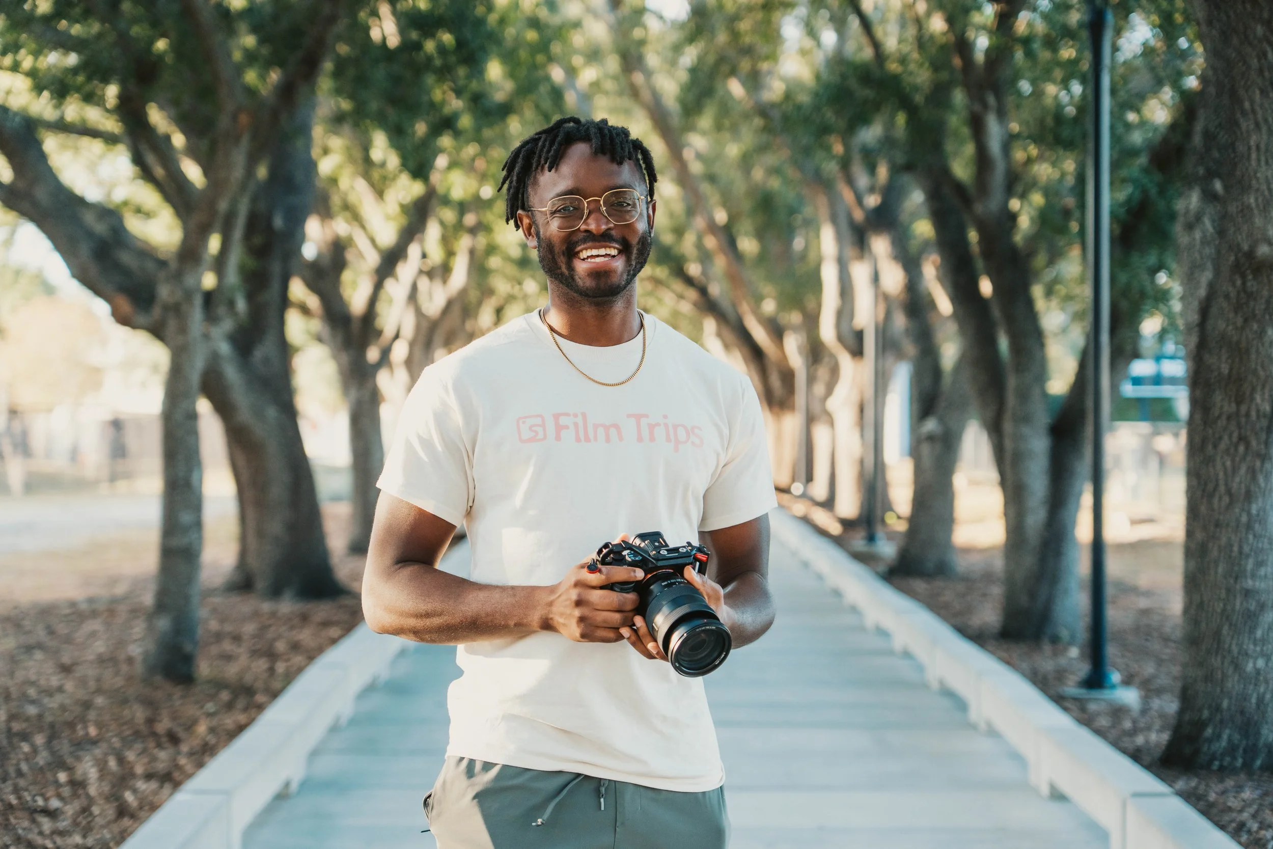 A smiling man holding a camera standing on a sidewalk lined with trees.