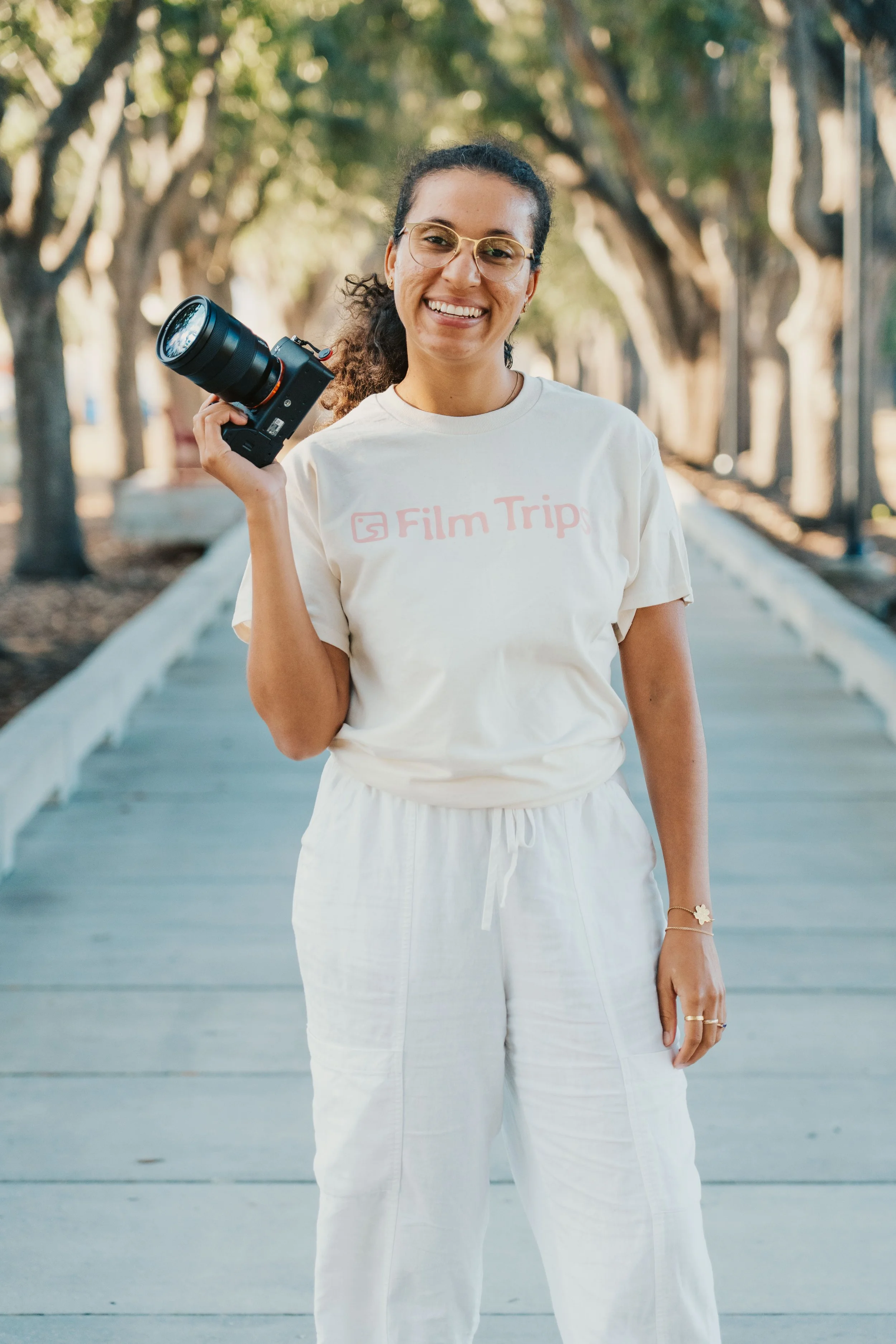 A woman smiling and holding a camera over her shoulder, standing on a wooden pathway lined with trees.