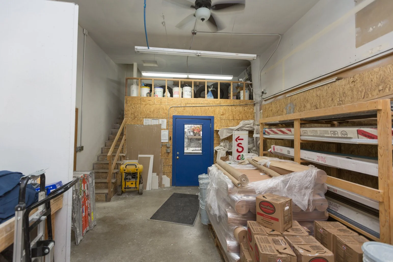 Interior view of Olbricht Drywall warehouse or store supplies area with construction materials, storage racks, a blue door, and stairs to an upper storage area.
