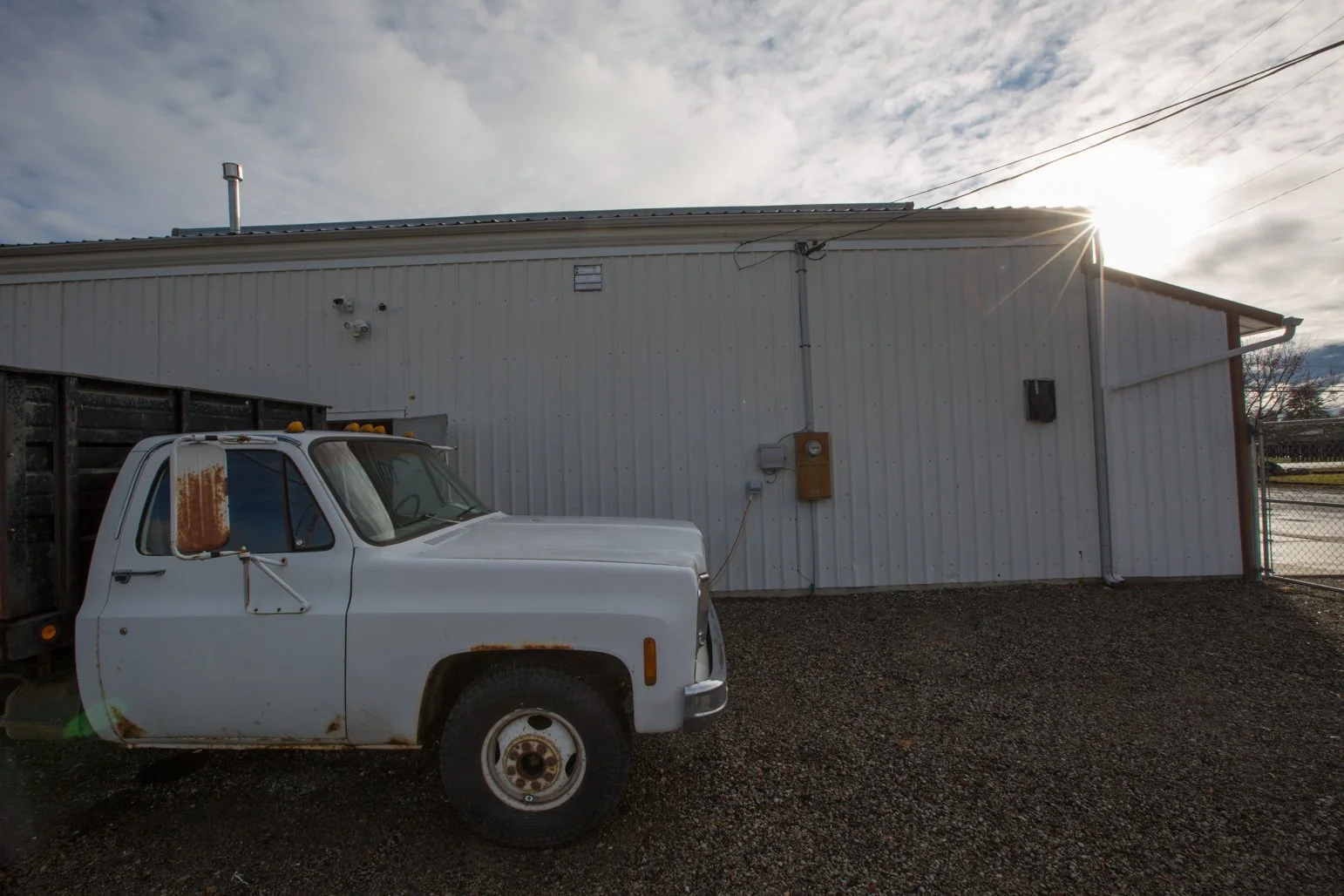 An old white truck parked outside Olbricht Drywall's building, sunlight visible behind the building, with wires and utility boxes on the wall.