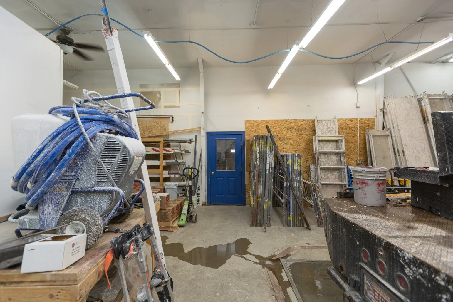 Interior view of Olbricht Drywall workshop or construction space with construction tools, ladders, a bucket, and construction materials, and a blue door at the back.