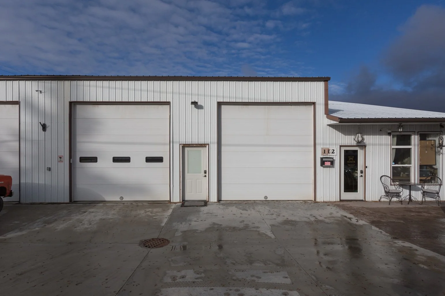Exterior of Olbricht Drywall's building with three garage doors,and a small seating area with a round table and two chairs, under a partly cloudy sky.