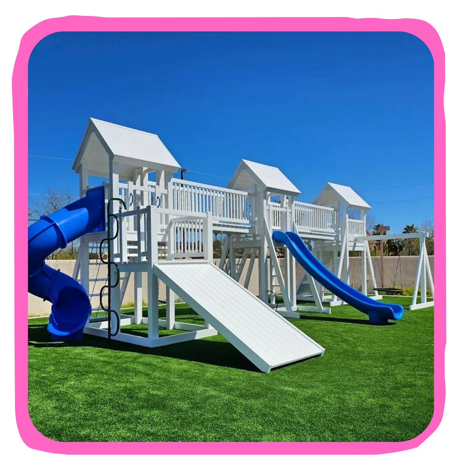 White wooden playground structure with blue slides, swings, and a climbing ramp on green grass under a clear blue sky.