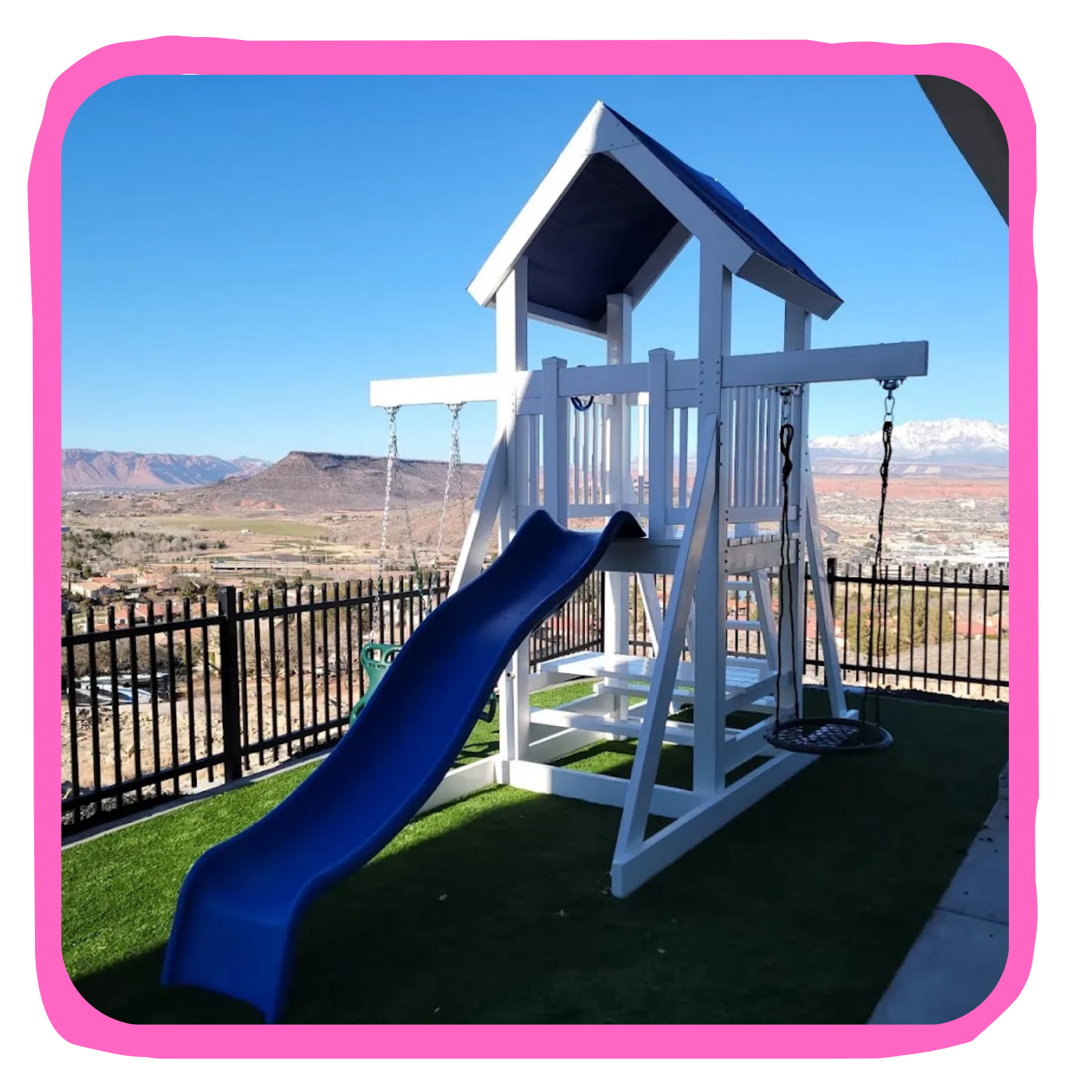 A children's backyard playset with a blue slide and swings on a grassy area, surrounded by a black fence, with a mountainous landscape in the background under a clear blue sky.