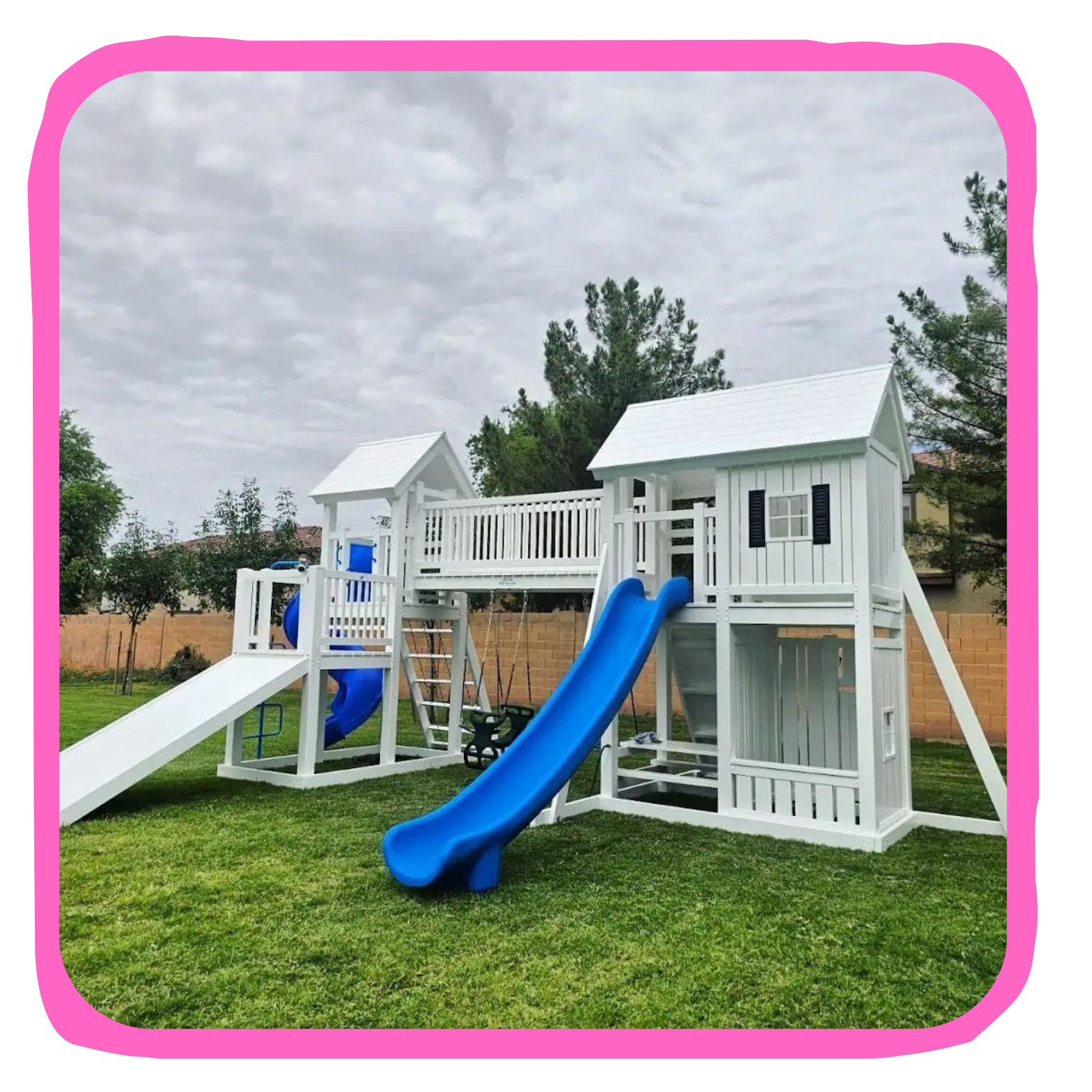 A white wooden outdoor playset with slides, swings, and a clubhouse, situated on a grassy backyard with trees and a brick wall in the background.