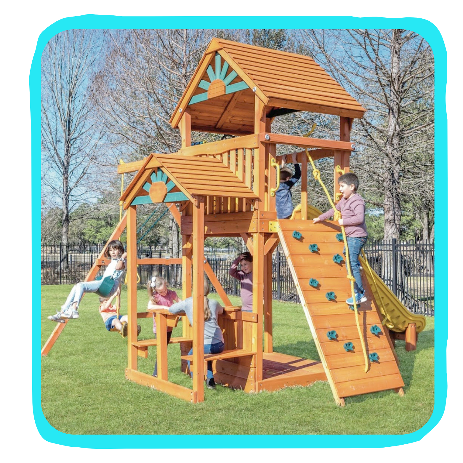 Children playing on a wooden multi-level playground structure with swings, a slide, and climbing features on a grassy field.