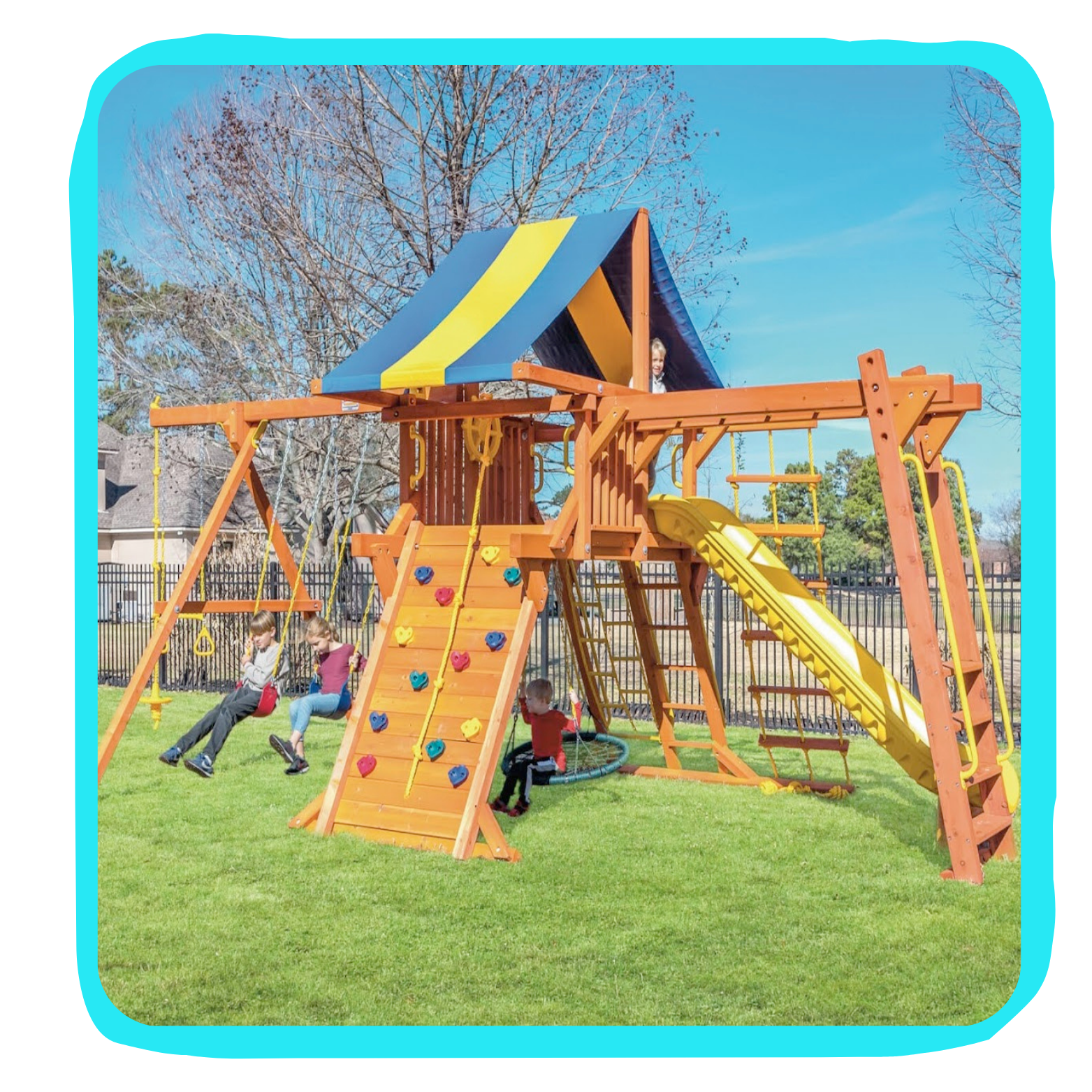 Children playing on a large wooden outdoor playset with swings, a slide, a climbing wall, and a covered platform in a grassy yard.