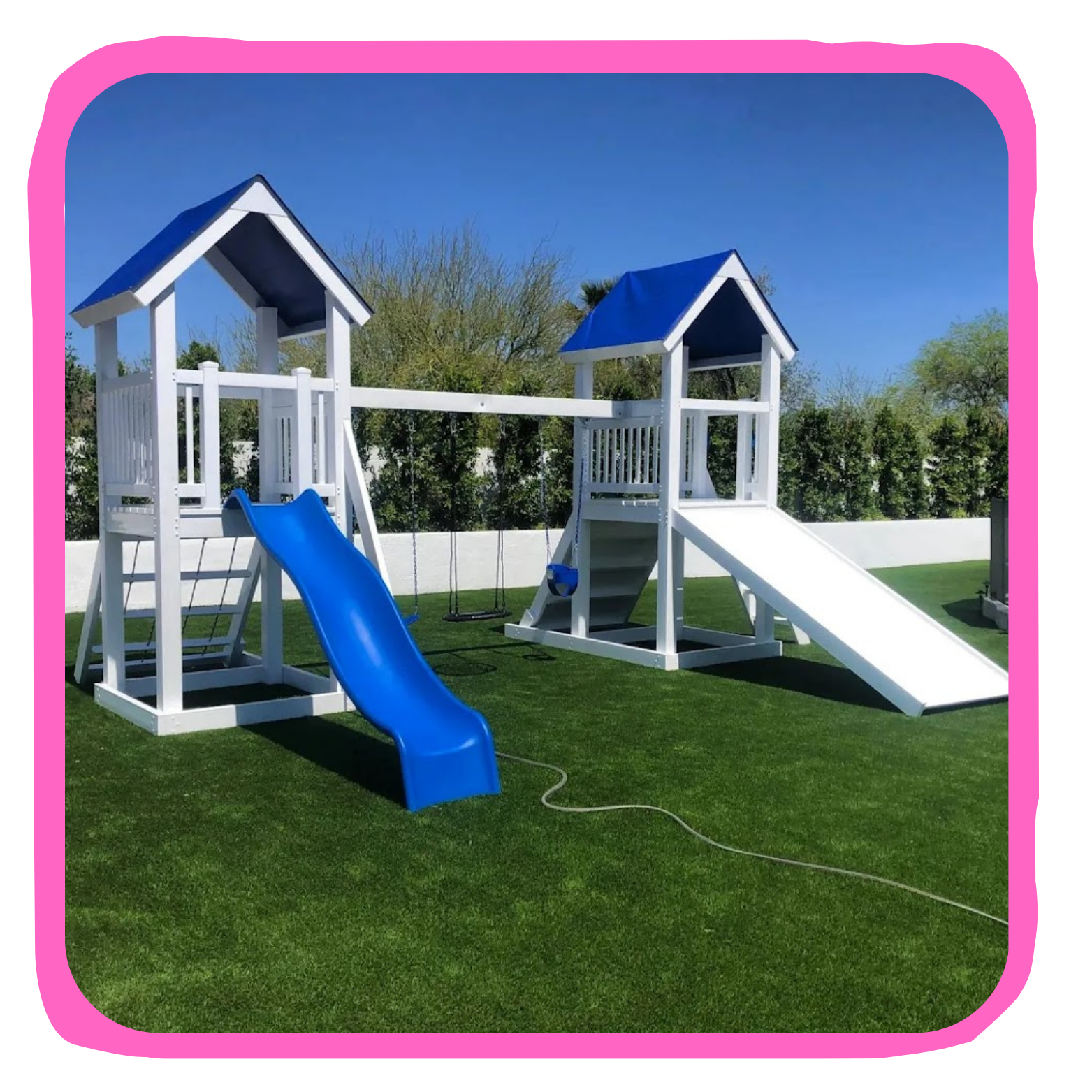 A white playground set with blue roofs, including a slide, swings, and climbing elements, on a grassy yard with a white wall and trees in the background.