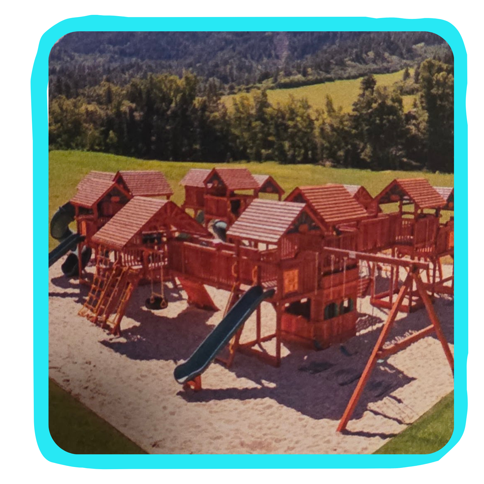 Wooden playground structure with slides, swings, and climbing areas situated on a bed of gravel with a grassy landscape and trees in the background.