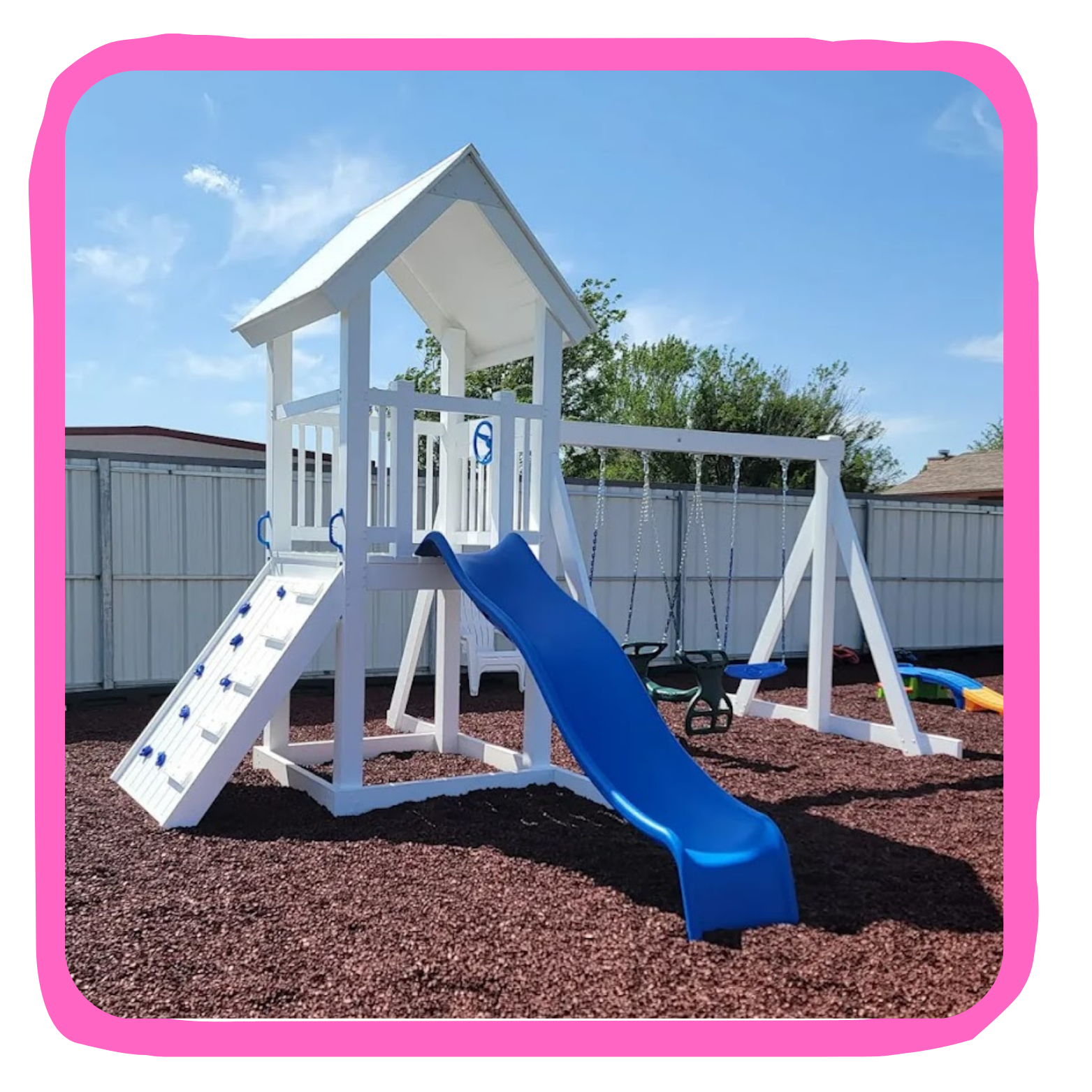 Children's outdoor playset with a slide, swings, and climbing wall on a yard with reddish mulch, fenced yard, trees, and blue sky.