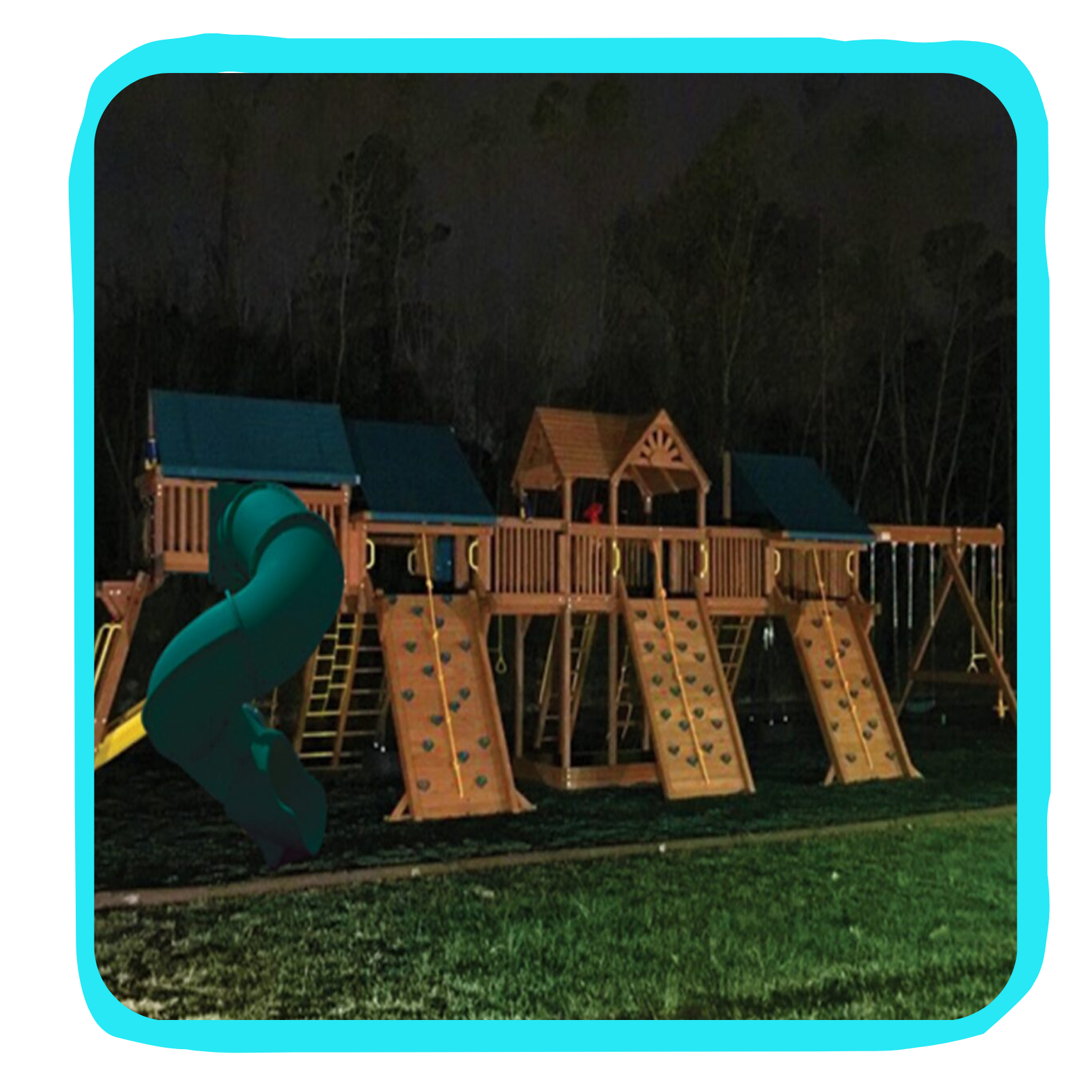 A wooden playground structure with slides, climbing walls, and swings illuminated at night, set against a background of dark trees.
