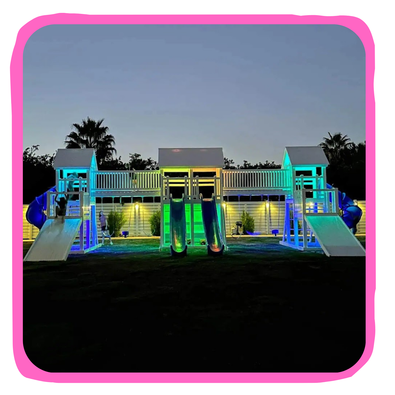 Outdoor playground structure illuminated with colorful lights against evening sky, with slides and climbing areas.