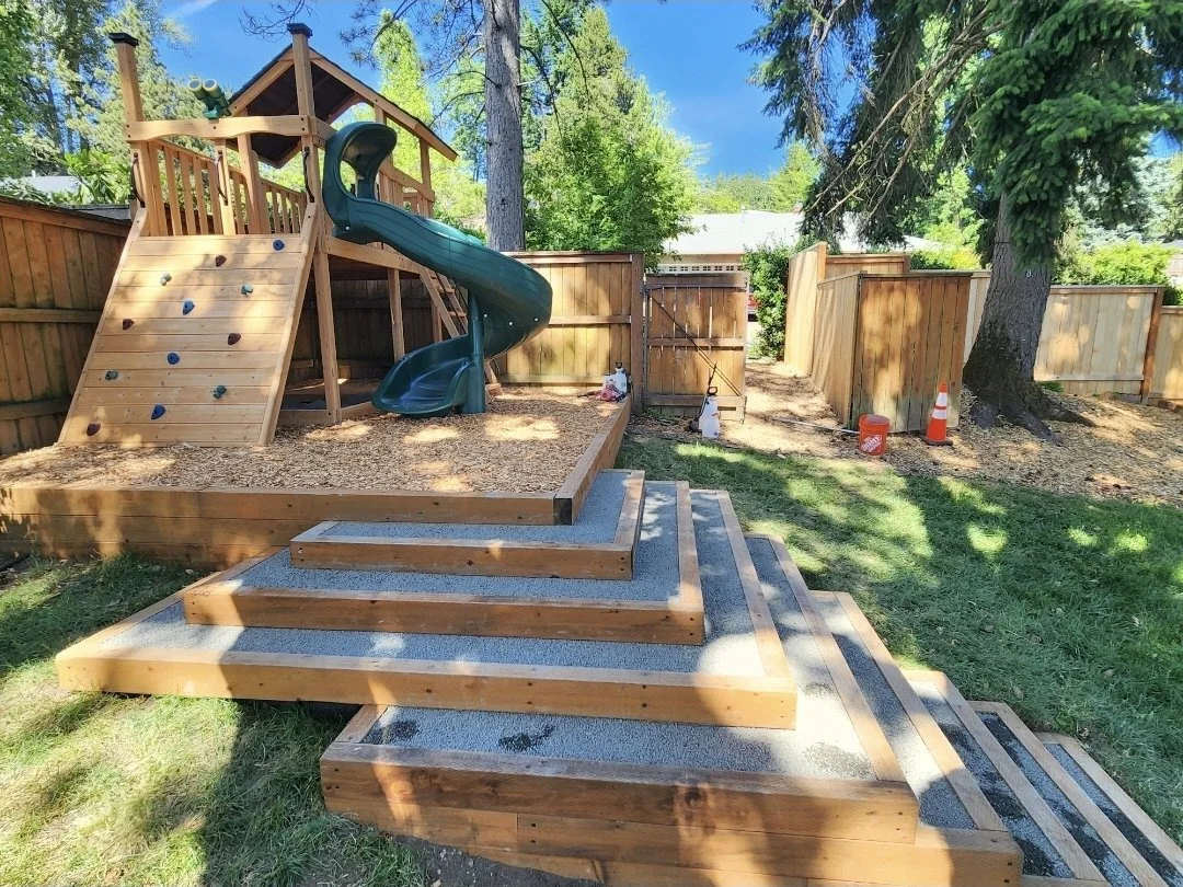 A backyard with a wooden playset featuring a slide and climbing wall, surrounded by a wooden fence, trees, and green grass.