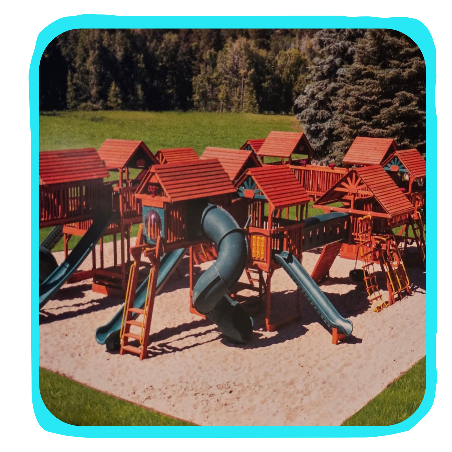 Colorful wooden playground with slides and climbing structures on sand surface, surrounded by green grass and trees.