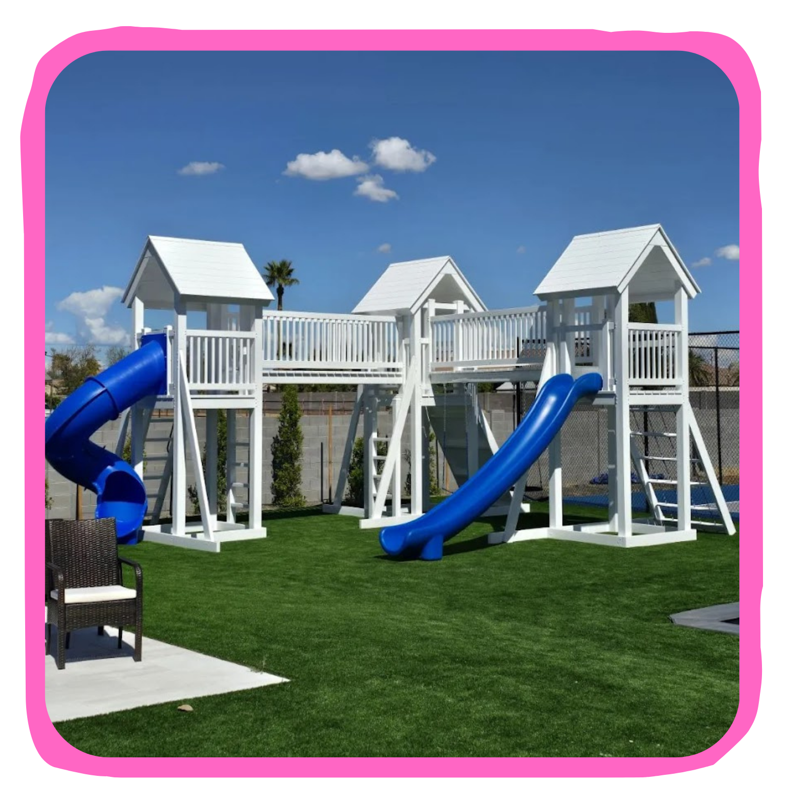 White wooden playground structure with blue slides on a green artificial lawn under a blue sky with a few clouds.