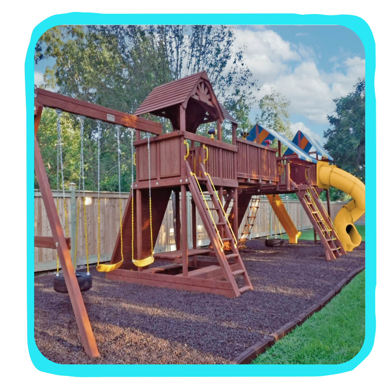 Playground set with swings, a slide, and a climbing ladder, surrounded by a wooden fence and a mulch ground cover.