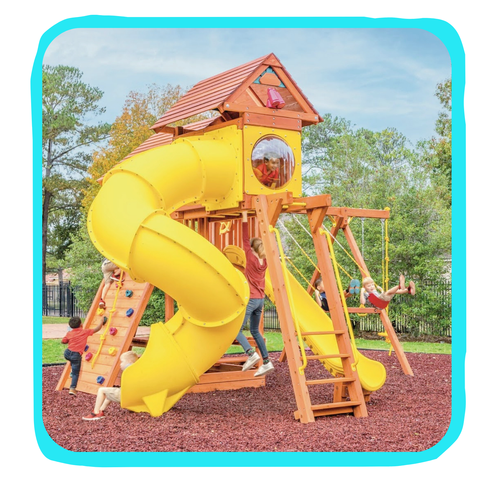 Children playing on a colorful wooden playground with a yellow slide and climbing wall, surrounded by trees and grass.