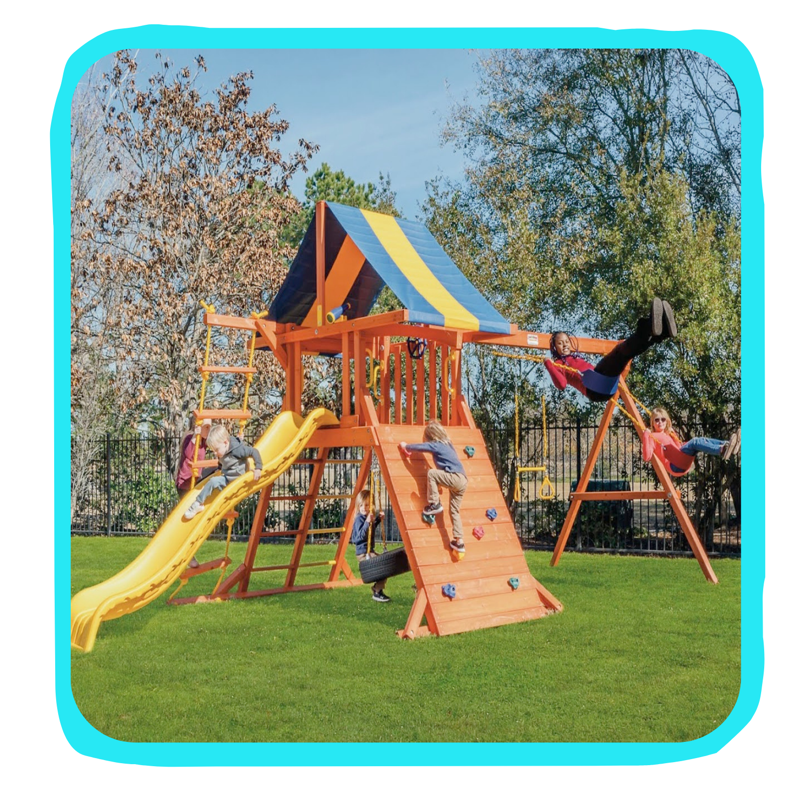 Children playing on outdoor playground structure with yellow slide, climbing wall, swing set, and small playhouse, surrounded by grass and trees.