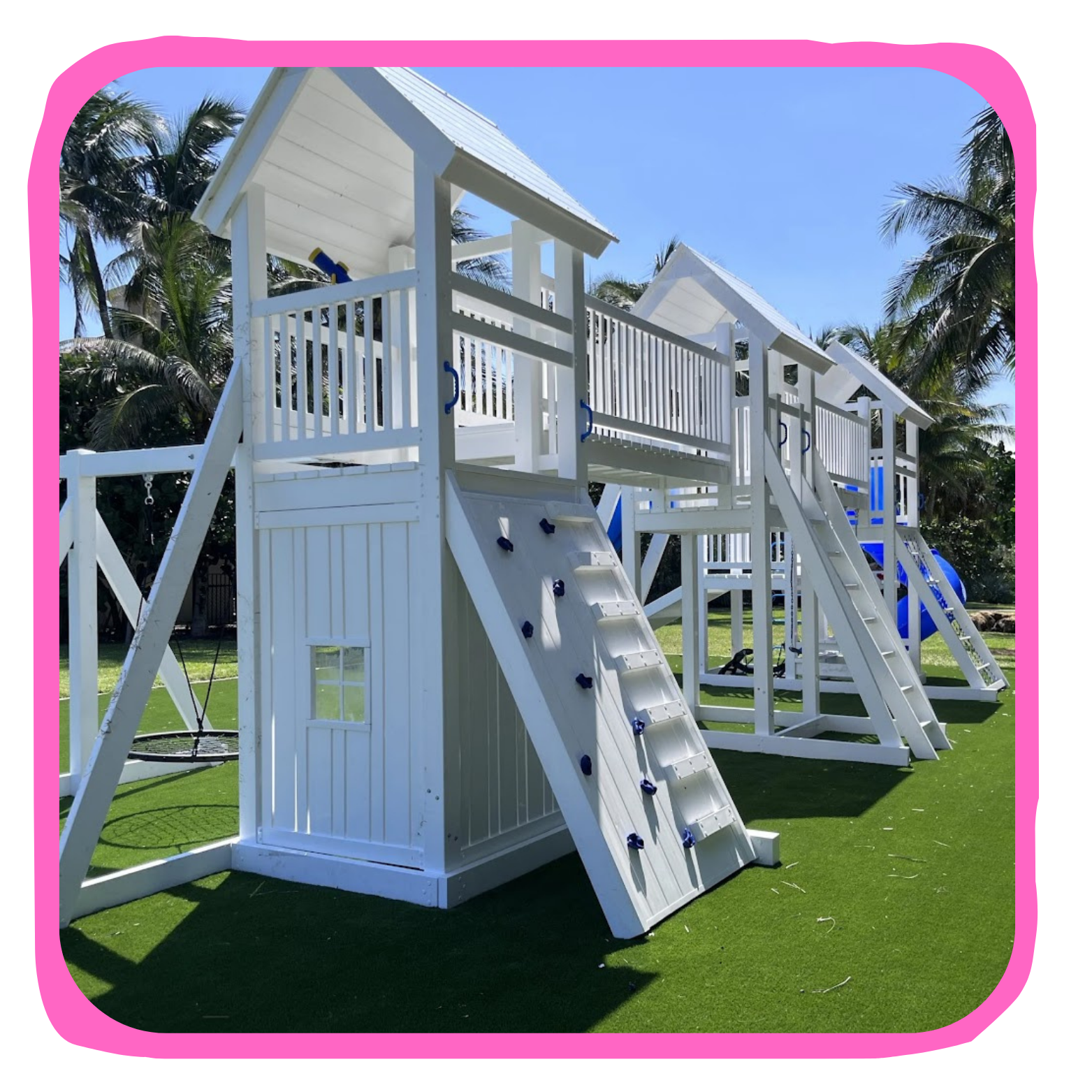 A white wooden playground structure with slides, a climbing wall, and a small house with a roof, set on artificial grass with palm trees and a clear blue sky in the background.